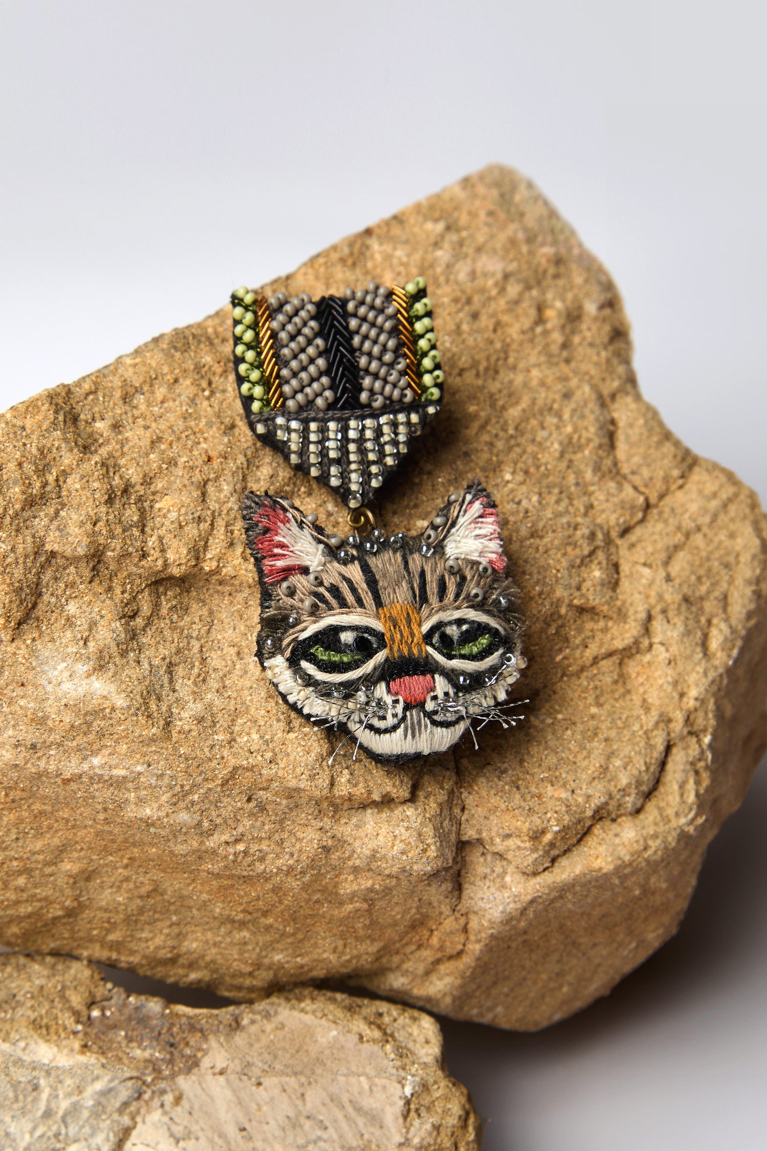 The Hand Embroidered Pussy Cat Brooch displays a cat's face, crafted with colorful threads and sparkling beads, set elegantly on a textured beige rock.