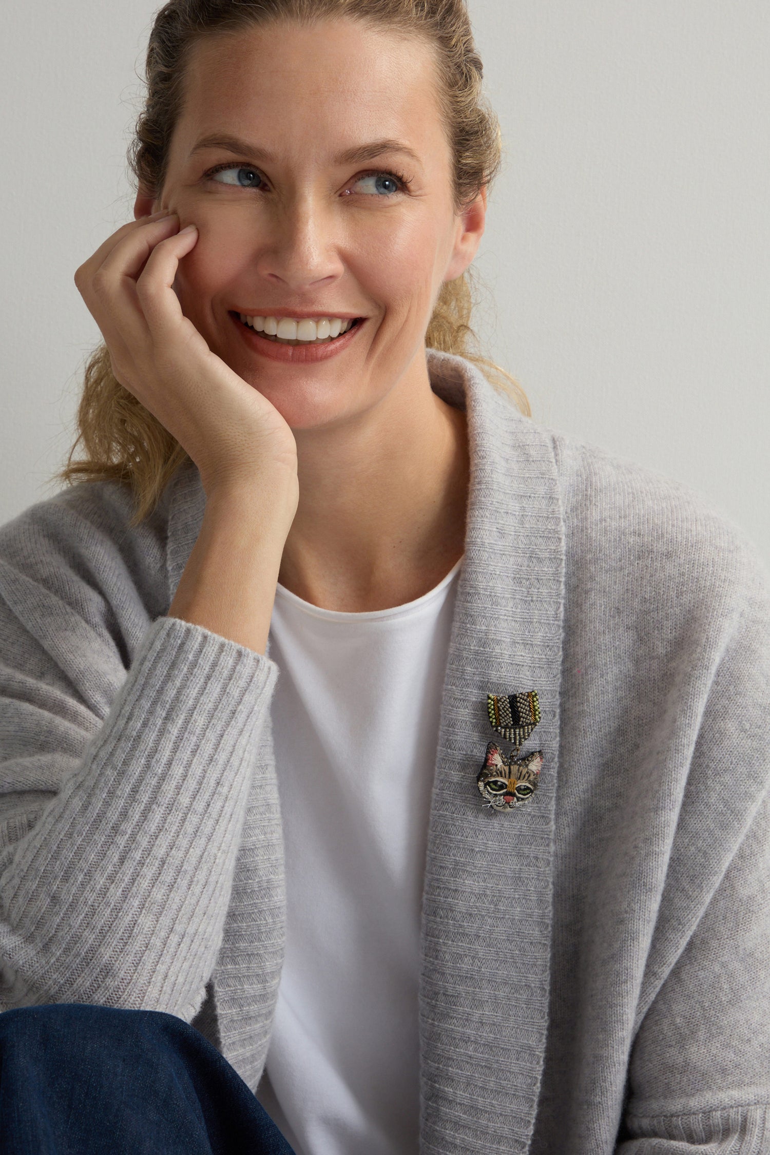 A smiling woman wears a gray cardigan and white shirt, highlighted by the Hand Embroidered Pussy Cat Brooch, adding a touch of charm.