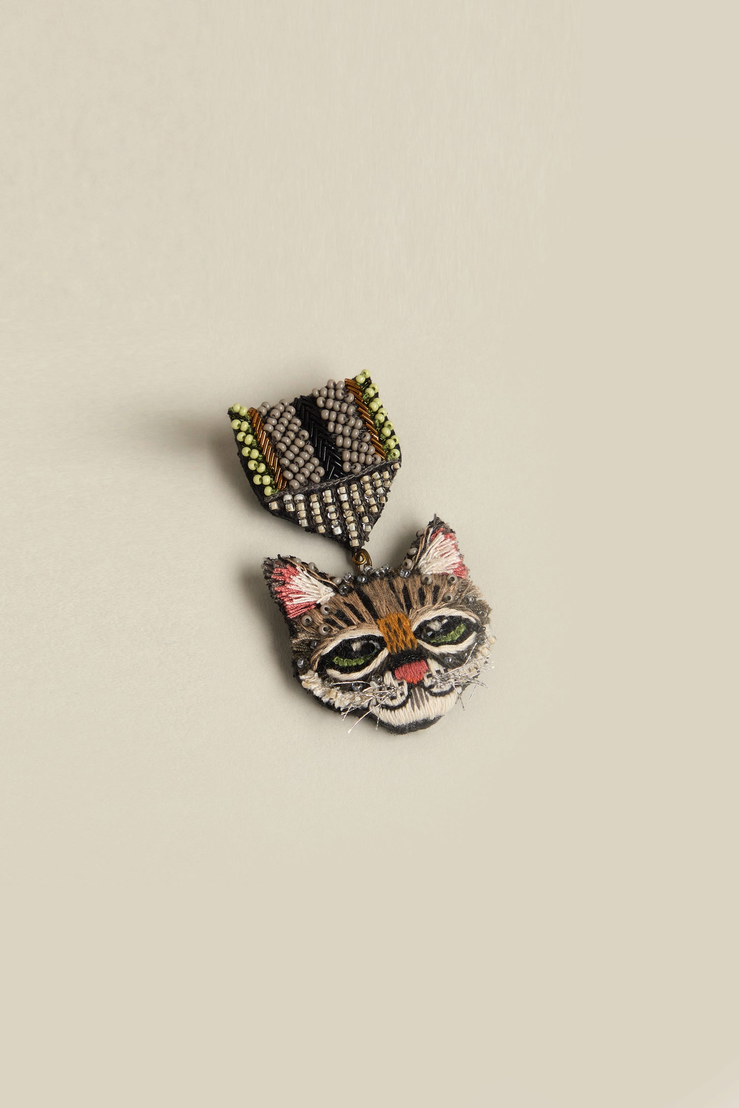 The Hand Embroidered Pussy Cat Brooch, with green eyes, pink ears, and brown fur, features an abstract beaded top on a plain background. It's a whimsical accessory that adds charm to any outfit.