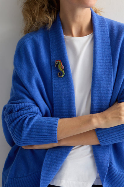 A person in a blue cardigan, arms crossed, and wearing a white t-shirt underneath is adorned with Trovelore's Hand Beaded Spotted Seahorse Brooch, featuring intricate embroidery and vibrant details.