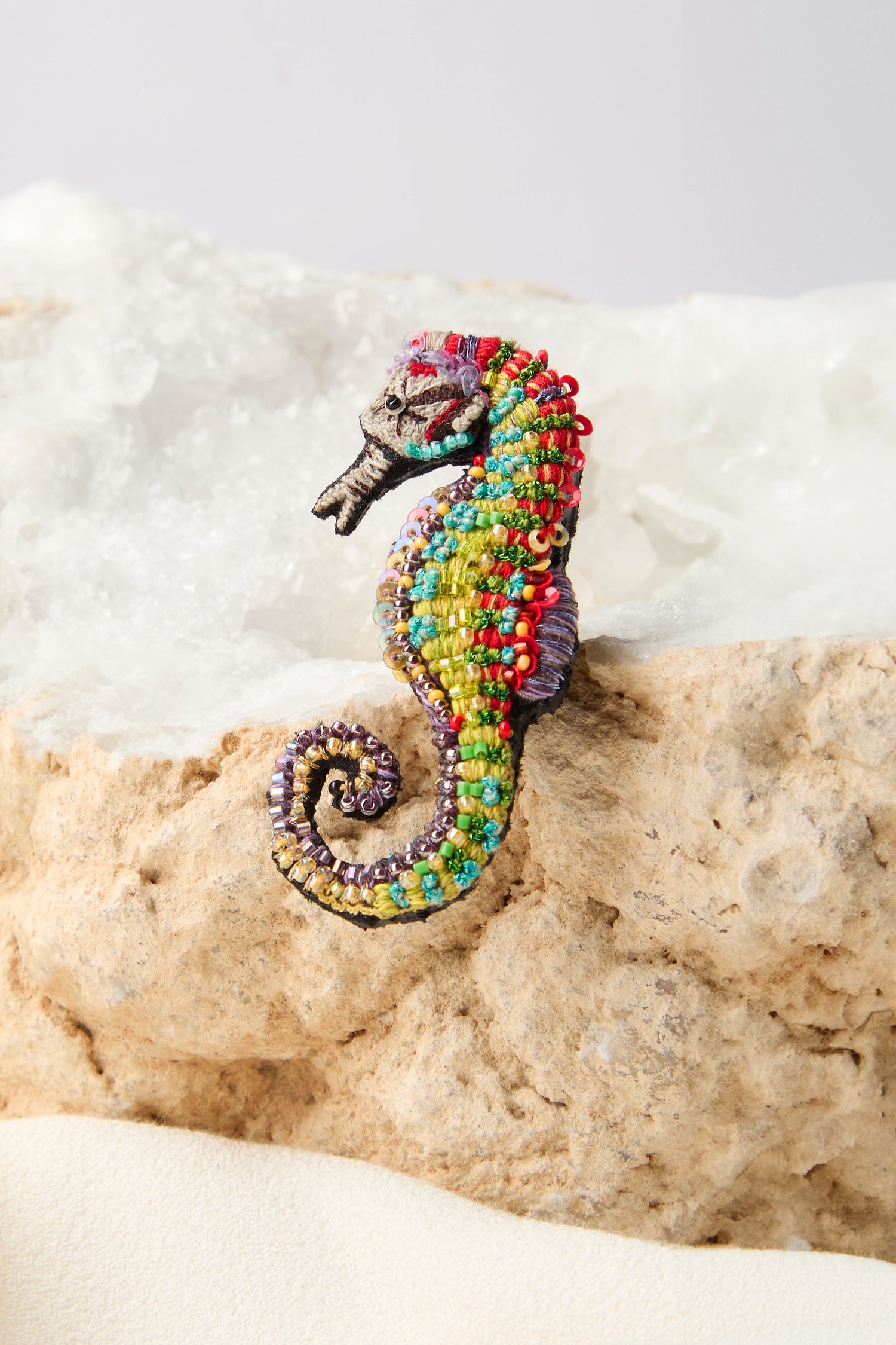 The Hand Beaded Spotted Seahorse Brooch, featuring vibrant colors and detailed embroidery, elegantly rests on a rock.