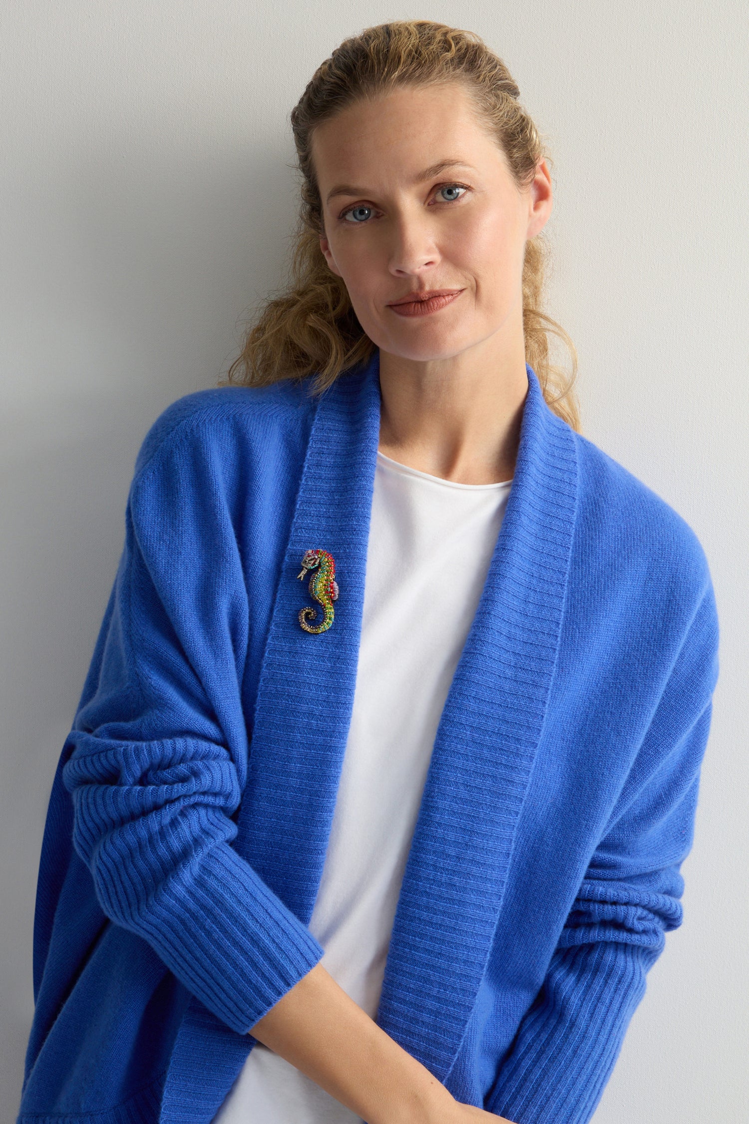 A calm-faced person wears a blue cardigan over a white shirt, elegantly accessorized with the Hand Beaded Spotted Seahorse Brooch. They stand against a plain background, highlighting the unique embroidery of their accessory.