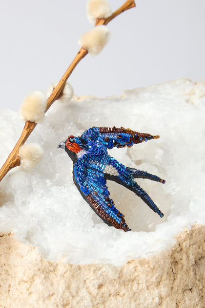 A Hand Beaded Flying Swallow Brooch lies gracefully on a rock beside a twig with soft buds, displaying exquisite sparkling beadwork.