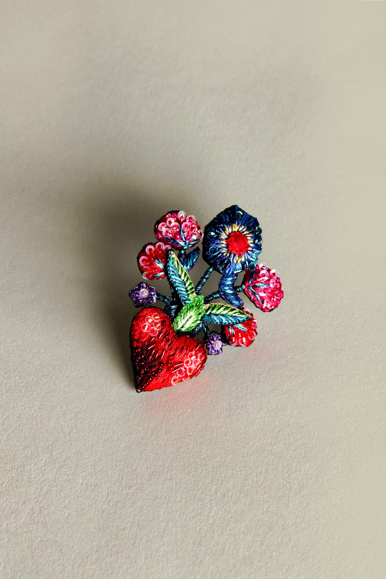 The Hand Beaded Flower Bouquet Brooch is an intricately crafted floral accessory with a red heart and colorful flowers on a neutral background, capturing elegance and detail reminiscent of Trovelore embroidery.