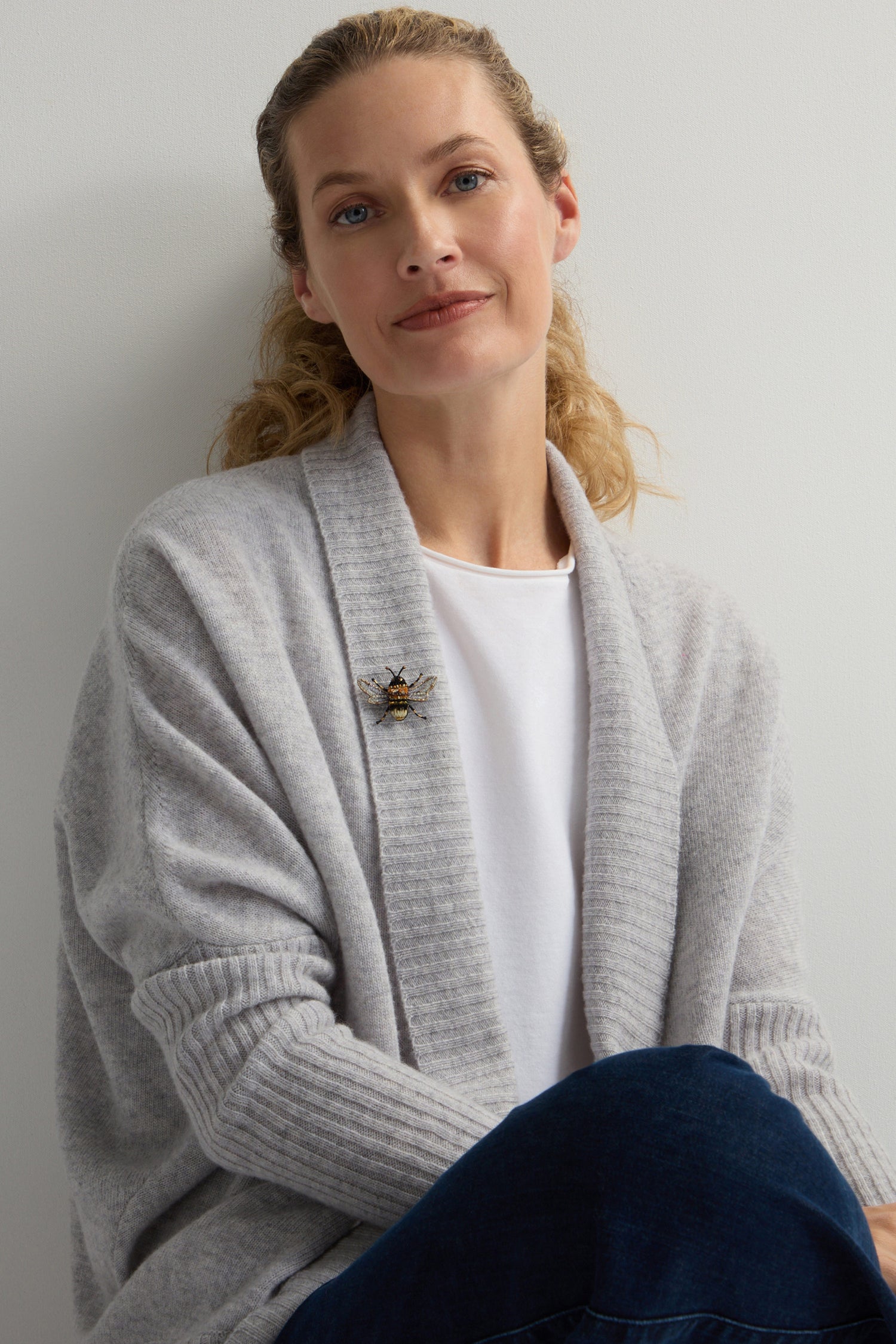 A blonde person in a gray cardigan, white shirt, and blue jeans sits against a light background, adorned with the Hand Beaded Humble Bee Brooch, highlighting dedication to sustainable fashion and intricate hand embroidery.