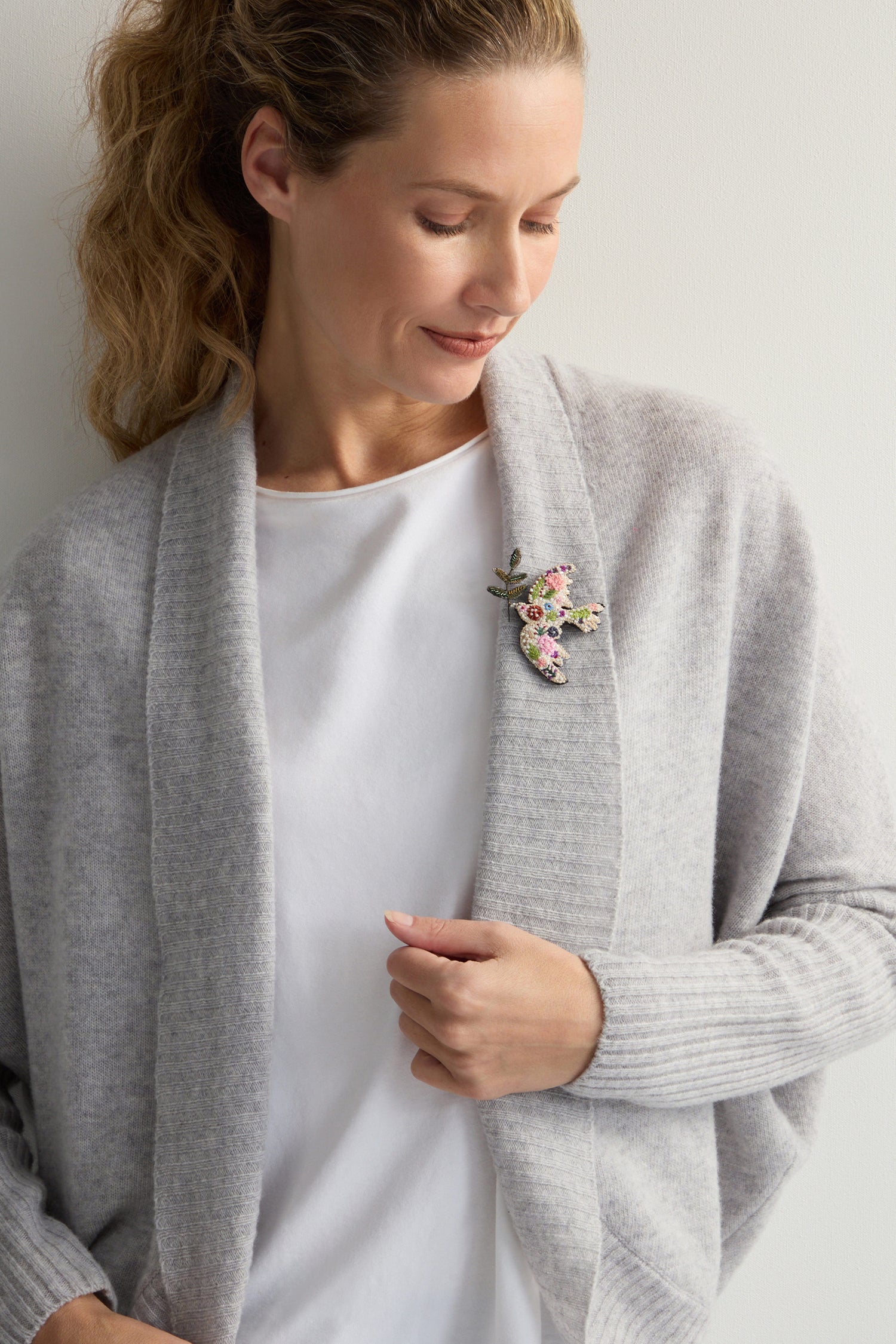 A woman in a light gray cardigan and white shirt looks down thoughtfully, wearing Trovelore's Hand Embroidered Peace Dove Brooch on her chest, elegantly capturing the essence of nature.