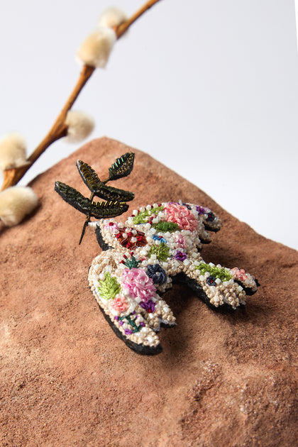 A colorful brooch, shaped like a bird with floral details, rests on a brown rock. Above it, a metallic dragonfly complements the scene. Crafted by Trovelore, this piece is reminiscent of the Hand Embroidered Peace Dove Brooch with intricate details.