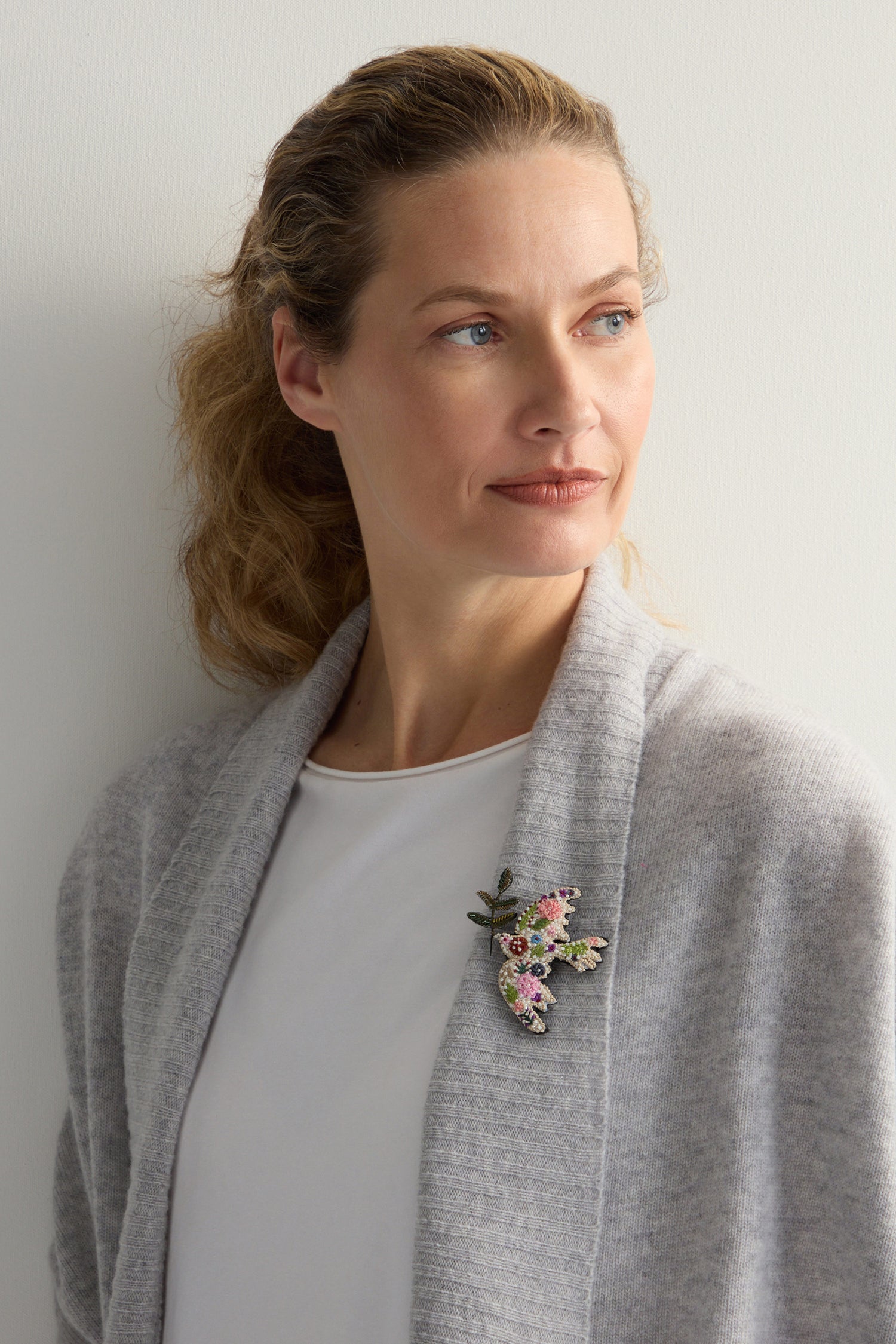 A woman in a gray cardigan and white shirt looks to the side, with a Hand Embroidered Peace Dove Brooch pinned on her cardigan, adding an elegant touch.