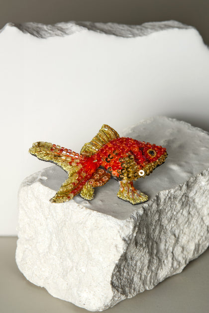 This Hand Beaded Fantail Goldfish Brooch, crafted with meticulous attention, embodies elegance through its red and gold beads on a textured white and gray stone surface.