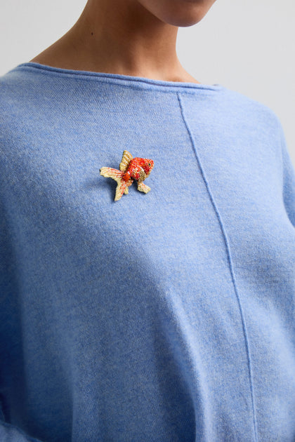 A person wearing a light blue sweater features a Trovelore handcrafted piece—a vibrant, Hand Beaded Fantail Goldfish Brooch—that adds a touch of whimsy to their ensemble.