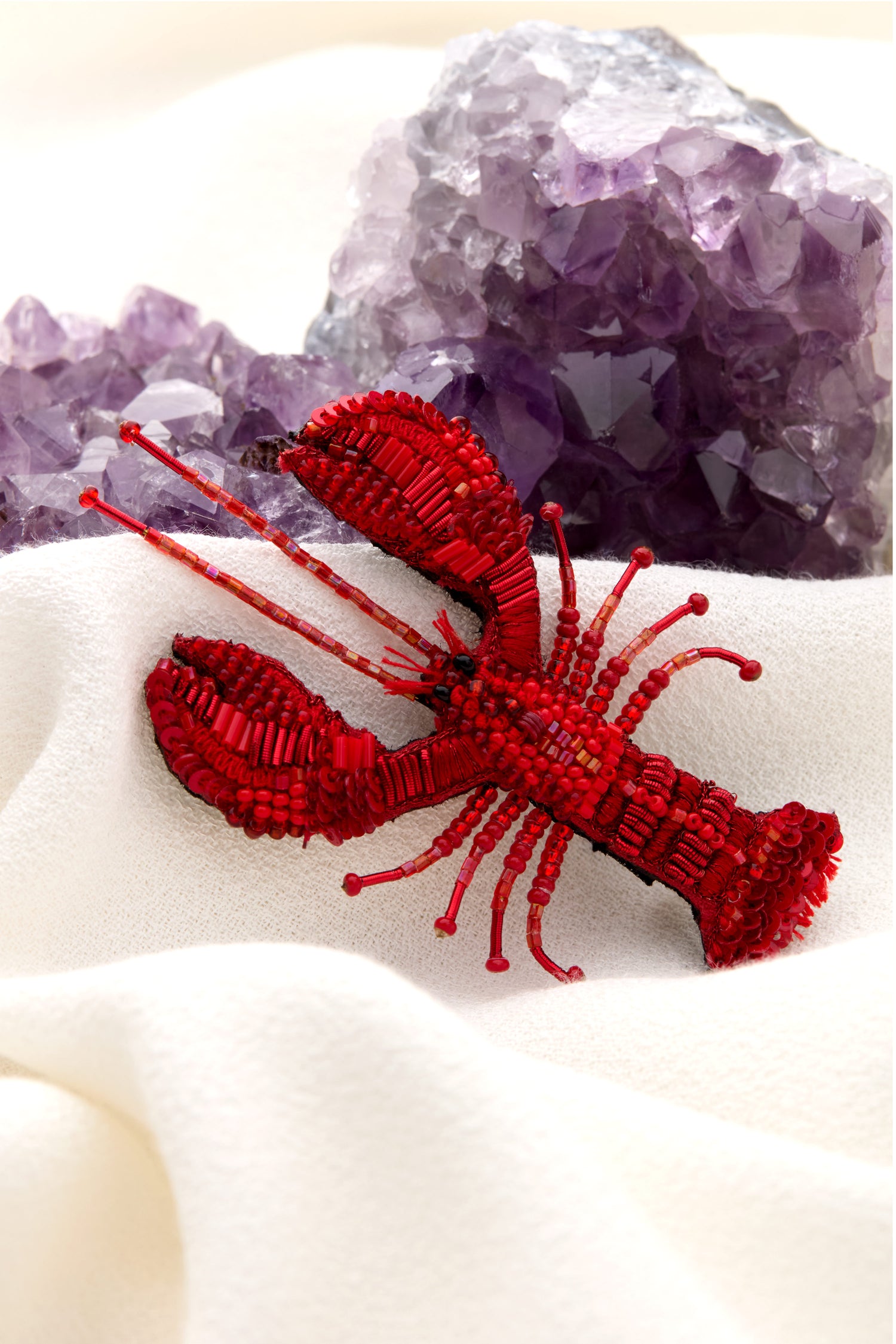 A Hand Embroidered Lobster Brooch from Trovelore, featuring intricate sequin and bead embellishments, sits on a white fabric background with purple crystal geodes in the distance.