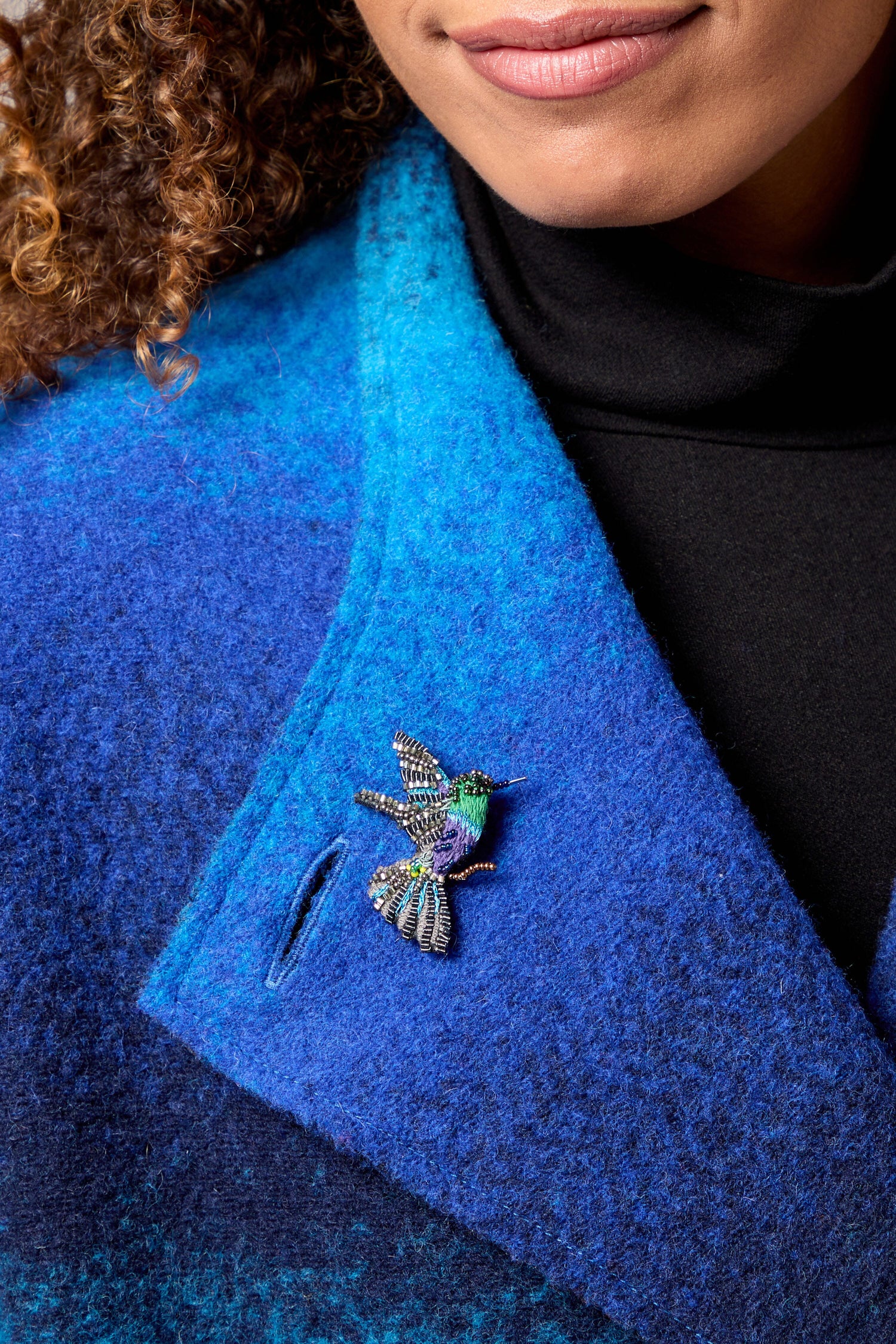 A woman wearing a blue coat with a Crowned Woodnymph Embroidered Brooch that sparkles.