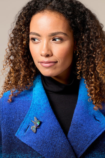 A woman wearing a blue coat with a Crowned Woodnymph Embroidered Brooch.