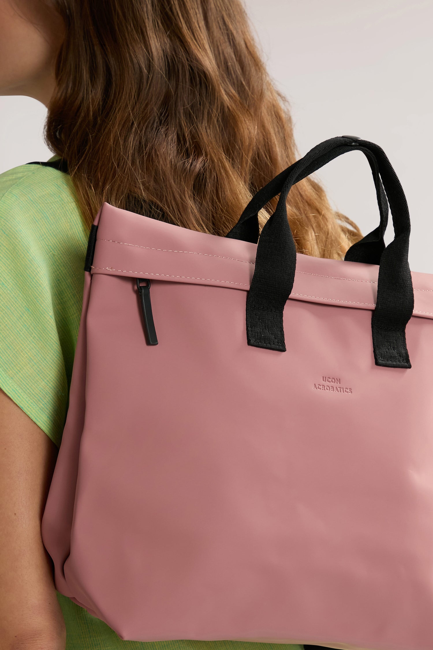 A person with long hair wearing a light green shirt carries a large pink Eliza Roll Top Backpack with black handles over their shoulder. The word "Ucon Acrobatics" is visible on the side of the bag, showcasing their commitment to sustainability, as it's made from recycled PET bottles.