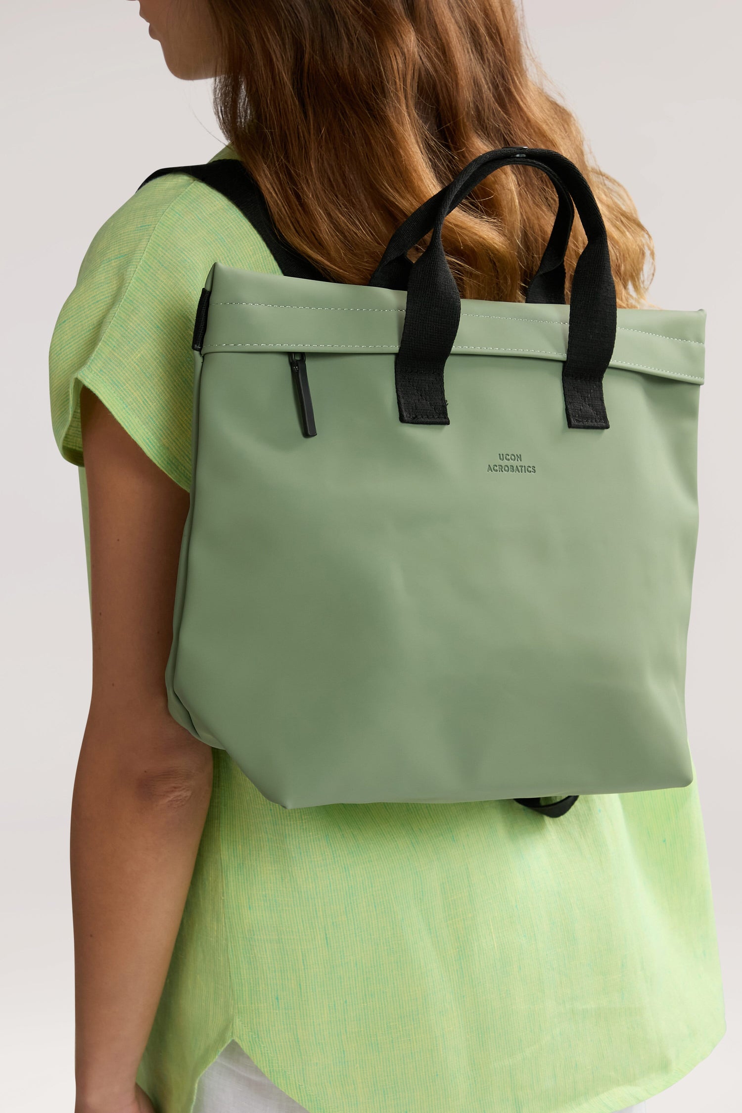 Person wearing a light green shirt and carrying an Eliza Roll Top Backpack with black handles and a zipper, made from recycled PET bottles, positioned like a backpack.