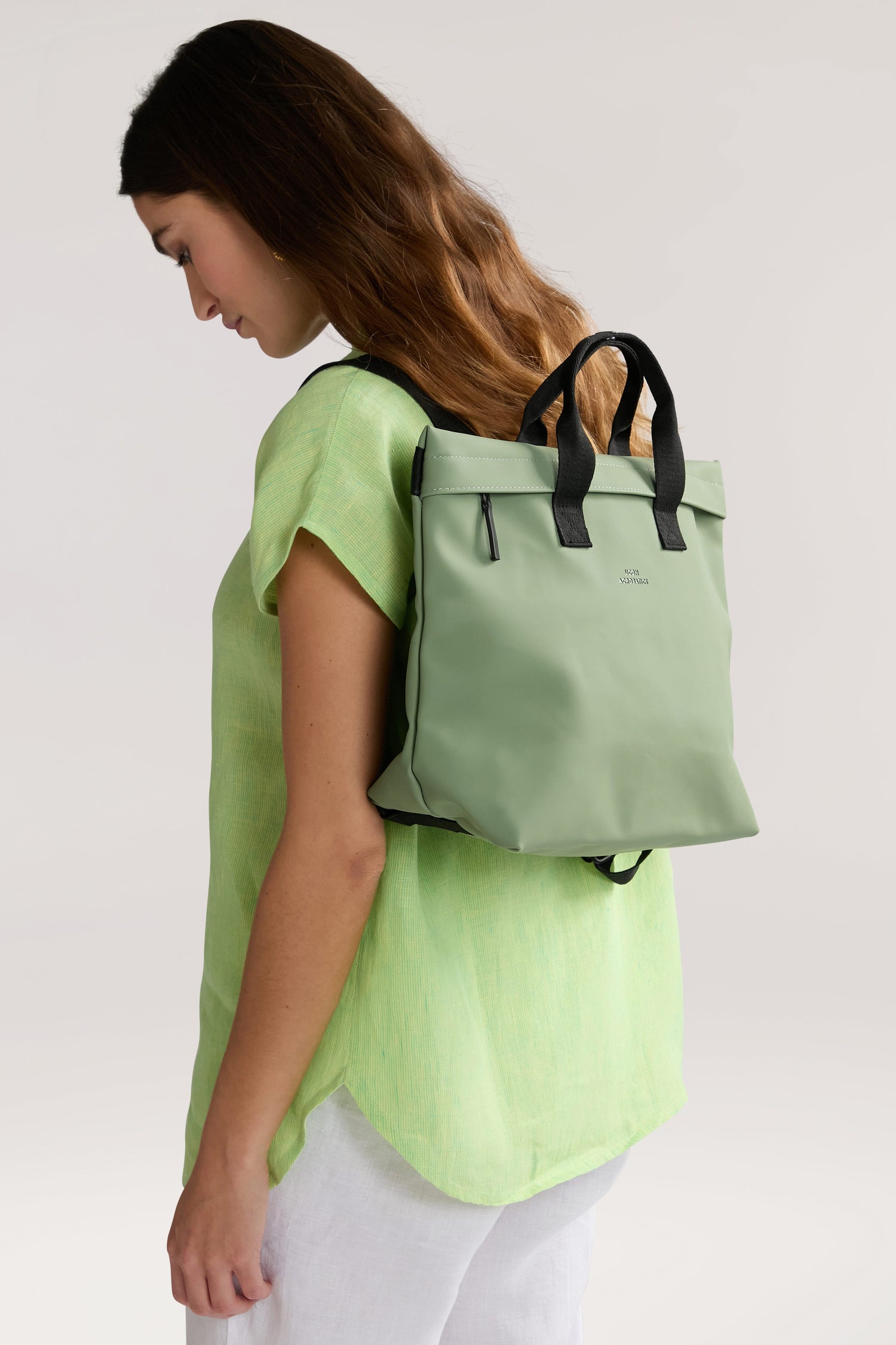 A woman wearing a light green top and white pants carries an Eliza Roll Top Backpack on her back. The light green backpack with black straps complements her outfit as she faces slightly sideways, looking downwards.