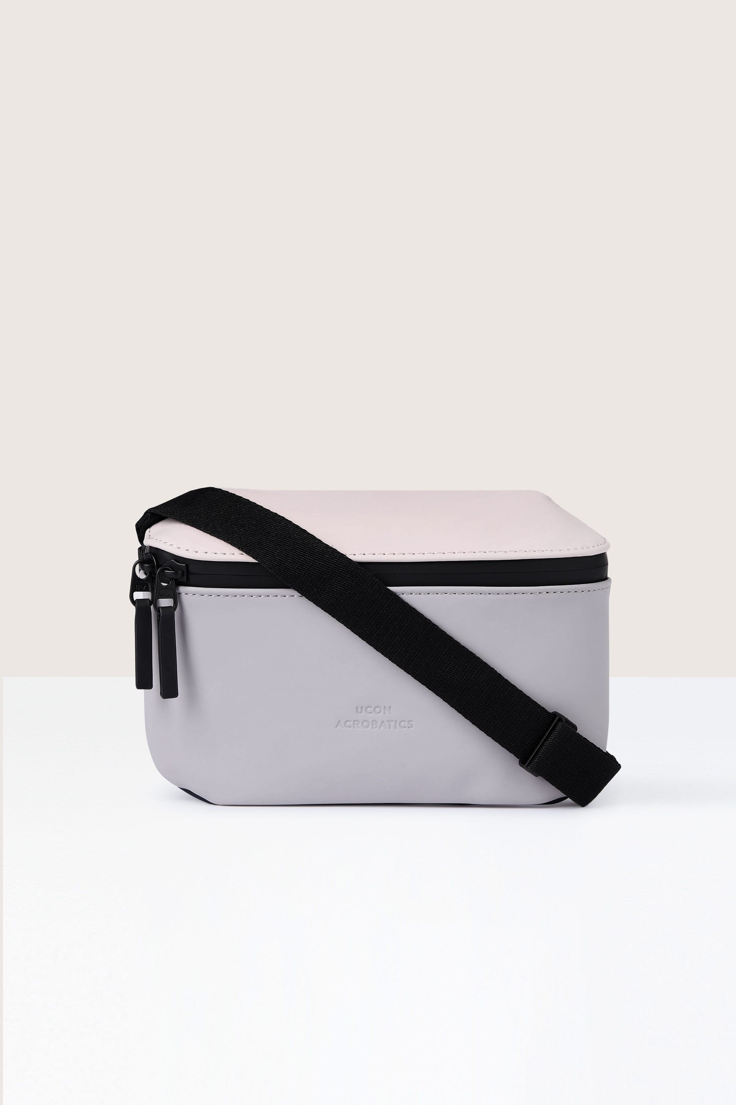 Introducing the Jona Crossbody Bag—a two-tone, rectangular shoulder bag made from water-repellent, vegan tech material. It features a black adjustable strap, a grey lower half, and a light pink upper half with a secure zipper closure.