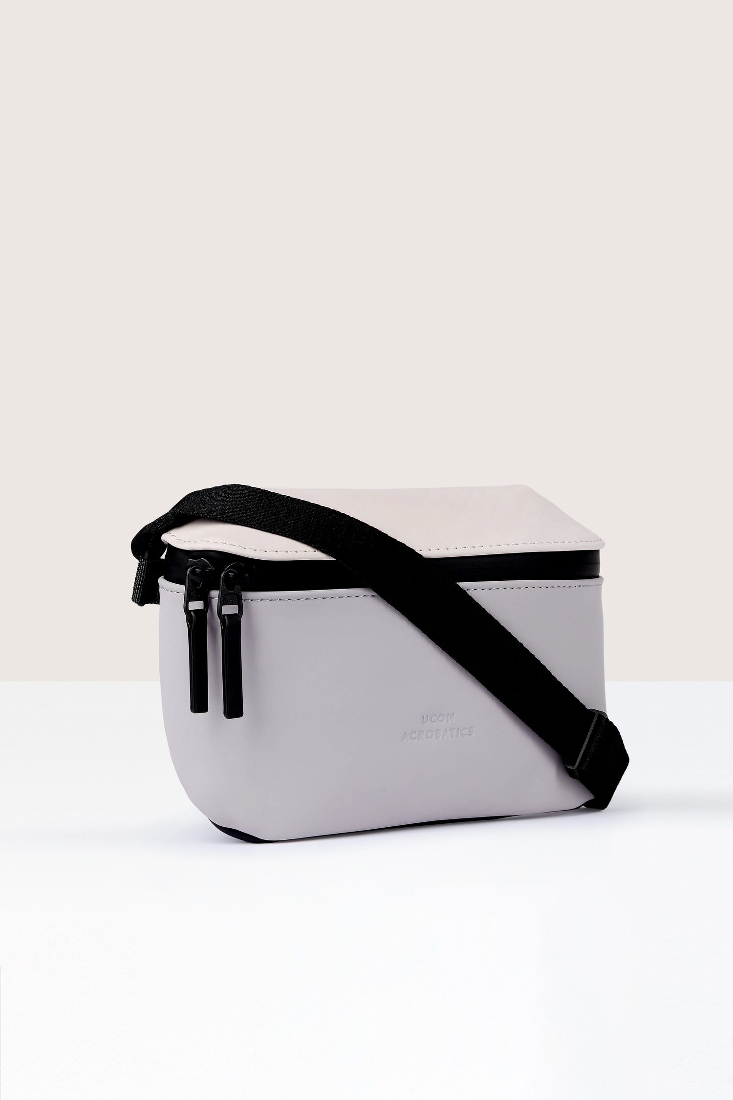 A small, light grey Jona Crossbody Bag with black zippers and an adjustable black strap, crafted from vegan tech material, placed on a white surface.