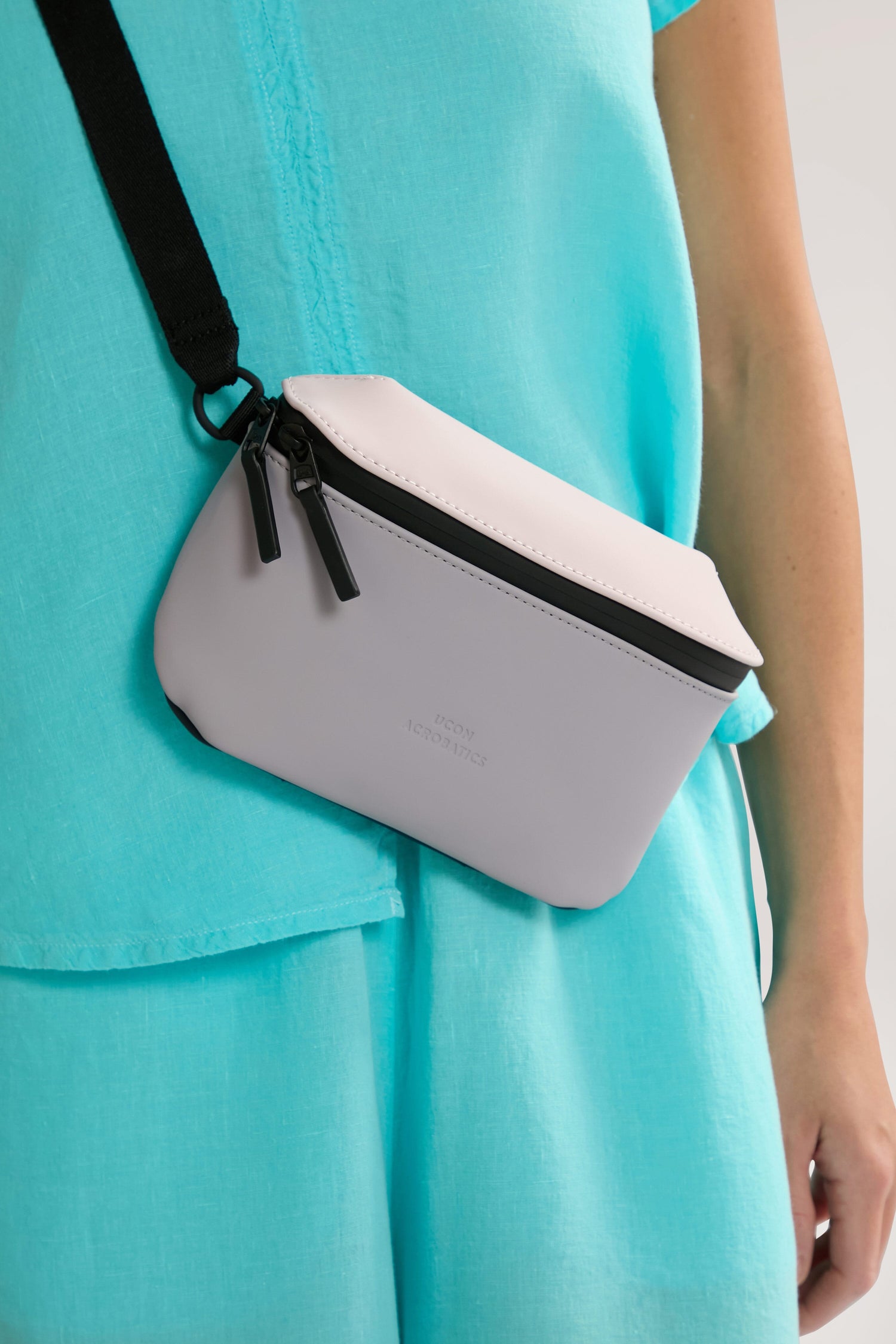 Person wearing a light blue outfit with a Jona Crossbody Bag made from vegan tech material, featuring black zippers and a black strap.