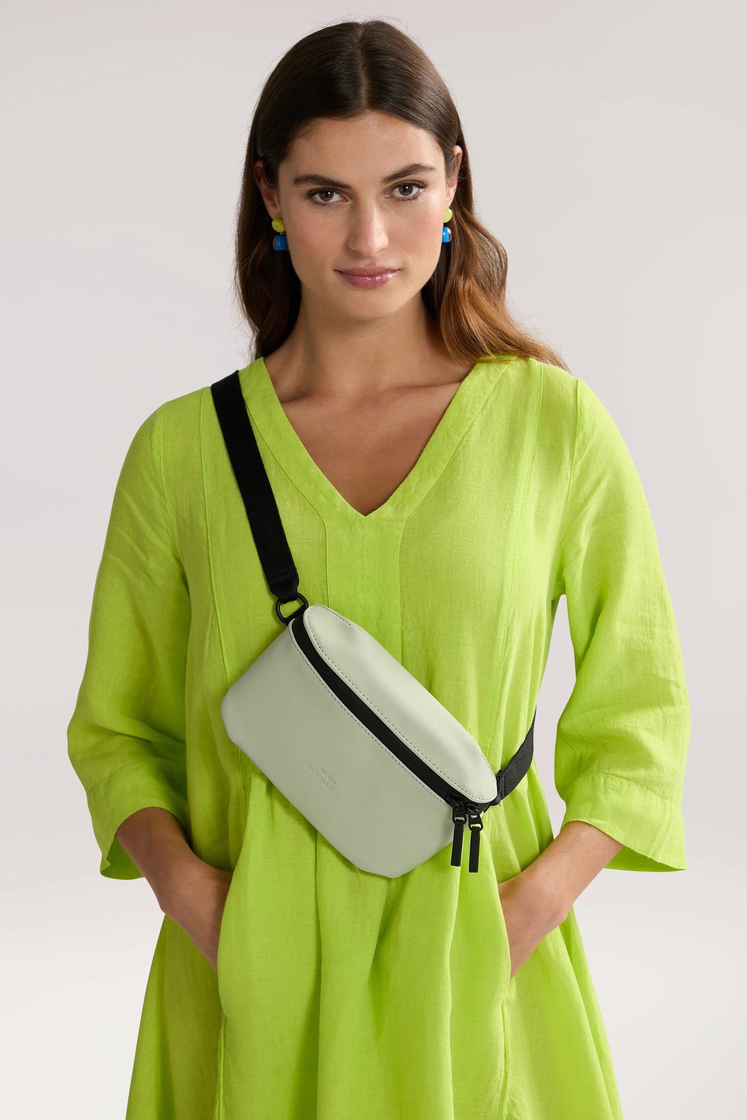 A woman is wearing a lime green dress and a light green Jona Crossbody Bag made from vegan tech material. She has her hands in the dress pockets and is looking directly at the camera.