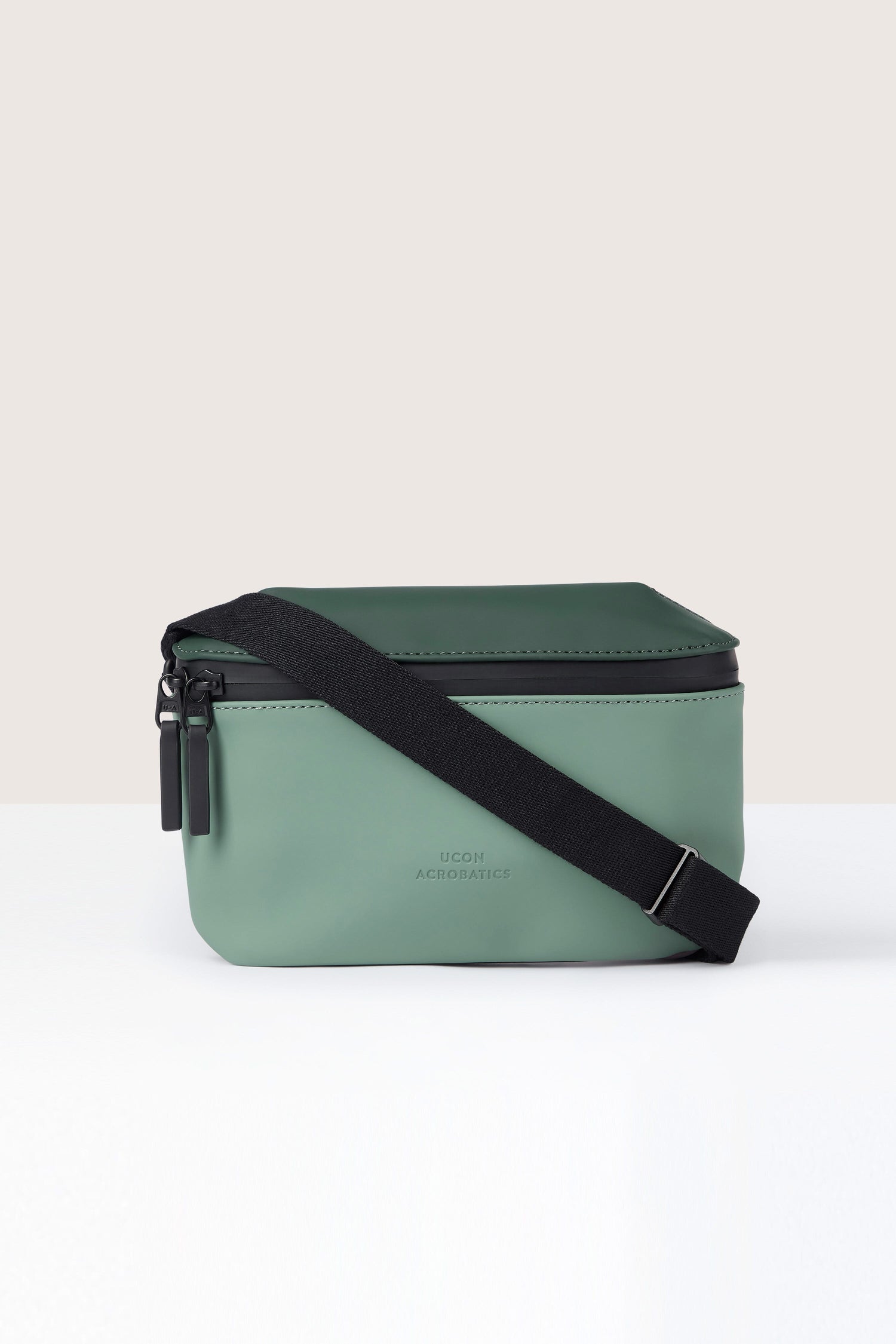 A water-repellent green Jona Crossbody Bag crafted from vegan tech material features a black adjustable strap and two black zipper pulls, all set against a neutral background.