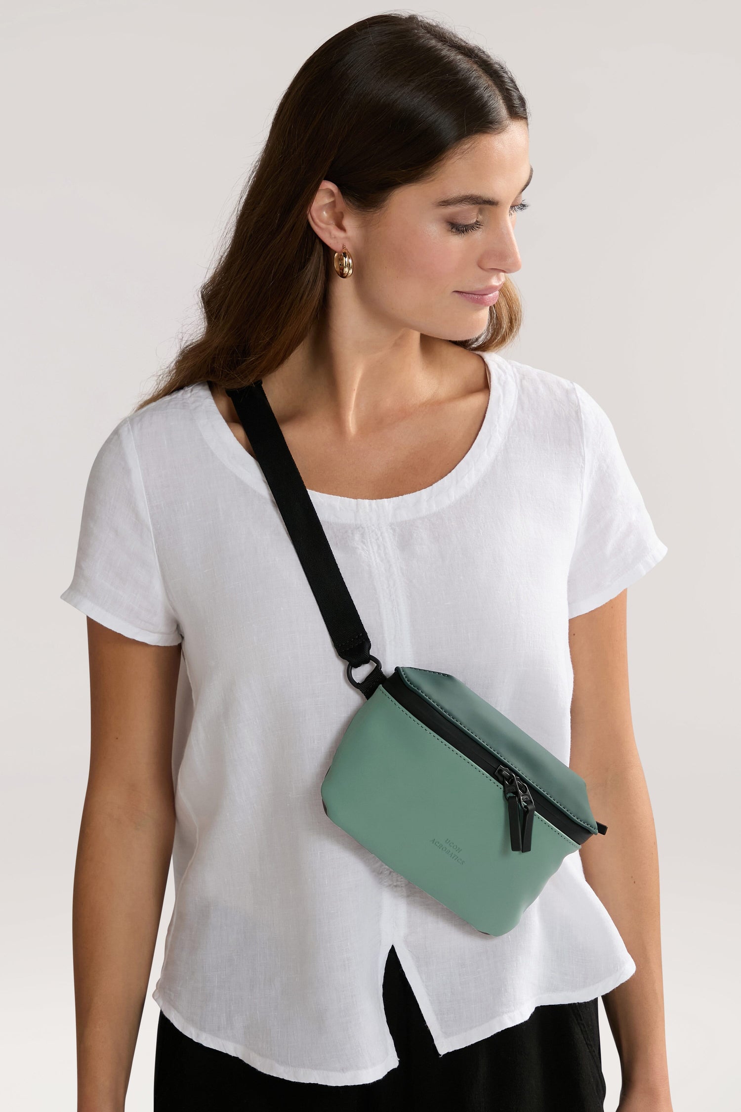 A woman wearing a white shirt and black pants is looking down and to the side. She has a Jona Crossbody Bag made of water-repellent vegan tech material strapped across her chest.