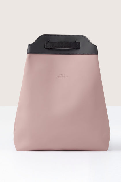 The Una Backpack features a minimalist pink bag with a black handle and top trim, crafted from recycled PET bottles, displayed against a neutral background.