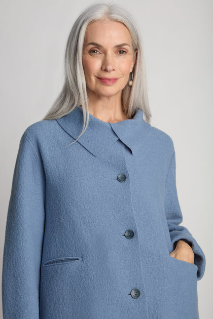 A woman with long grey hair is wearing a Collar Boiled Wool Jacket in blue, posing with one hand in her pocket and making a statement with the oversized fit of her outerwear.