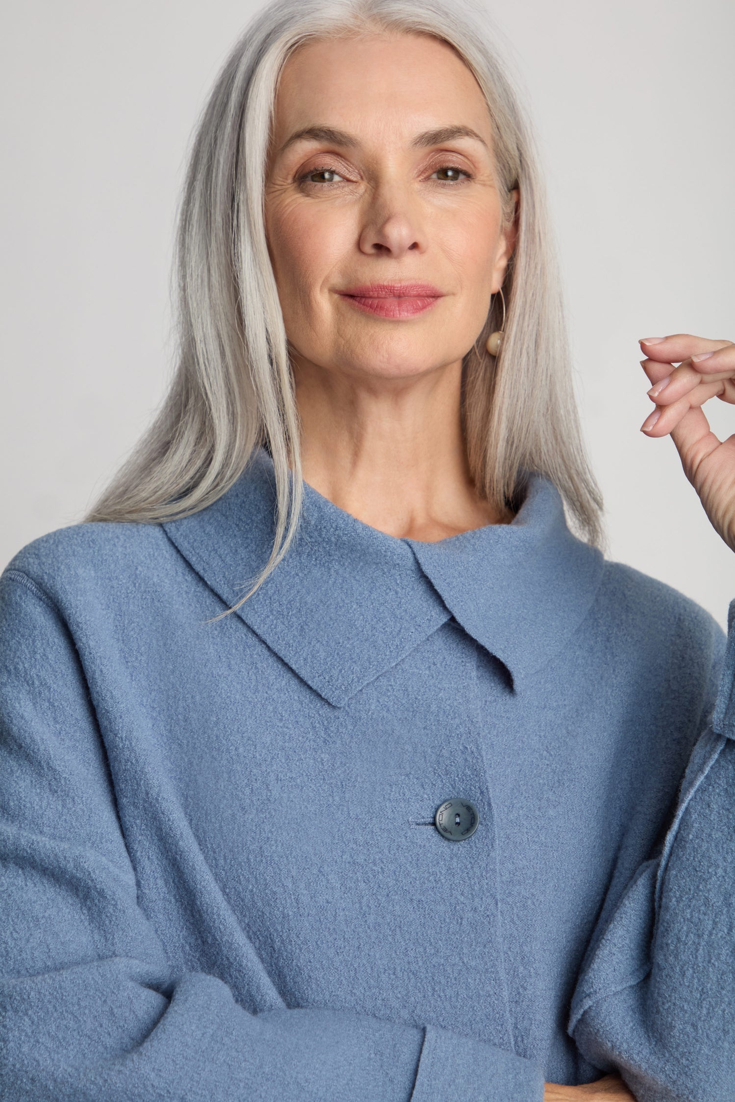 An older woman with long gray hair is wearing a Collar Boiled Wool Jacket in light blue, featuring a collar and a single visible button. She is looking at the camera with a slight smile, showcasing her statement outerwear in an oversized fit.