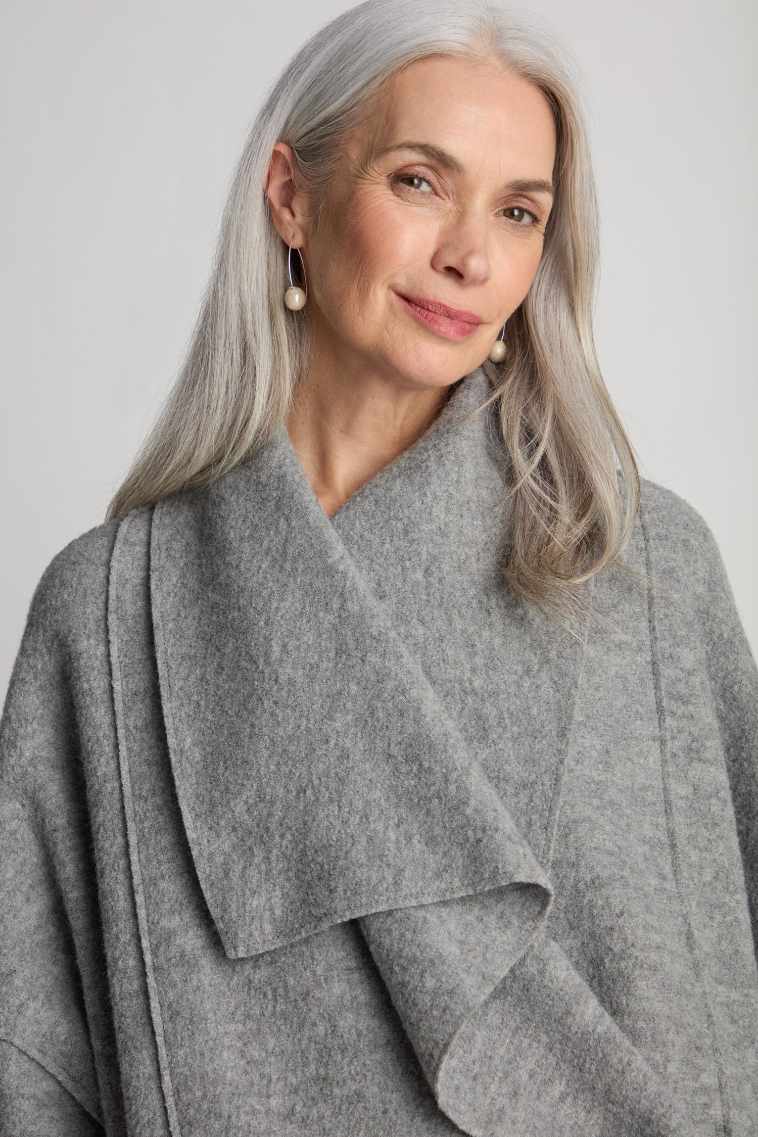 A woman with long gray hair, wearing the Shawl Collar Boiled Wool Coat with a safety pin closure and pearl earrings, smiles slightly against a neutral background.