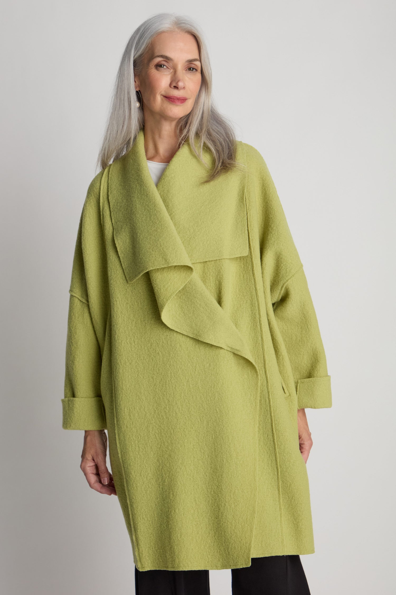 A person with long gray hair, smiling while standing against a plain background, is wearing a loose-fitting Shawl Collar Boiled Wool Coat in light green.