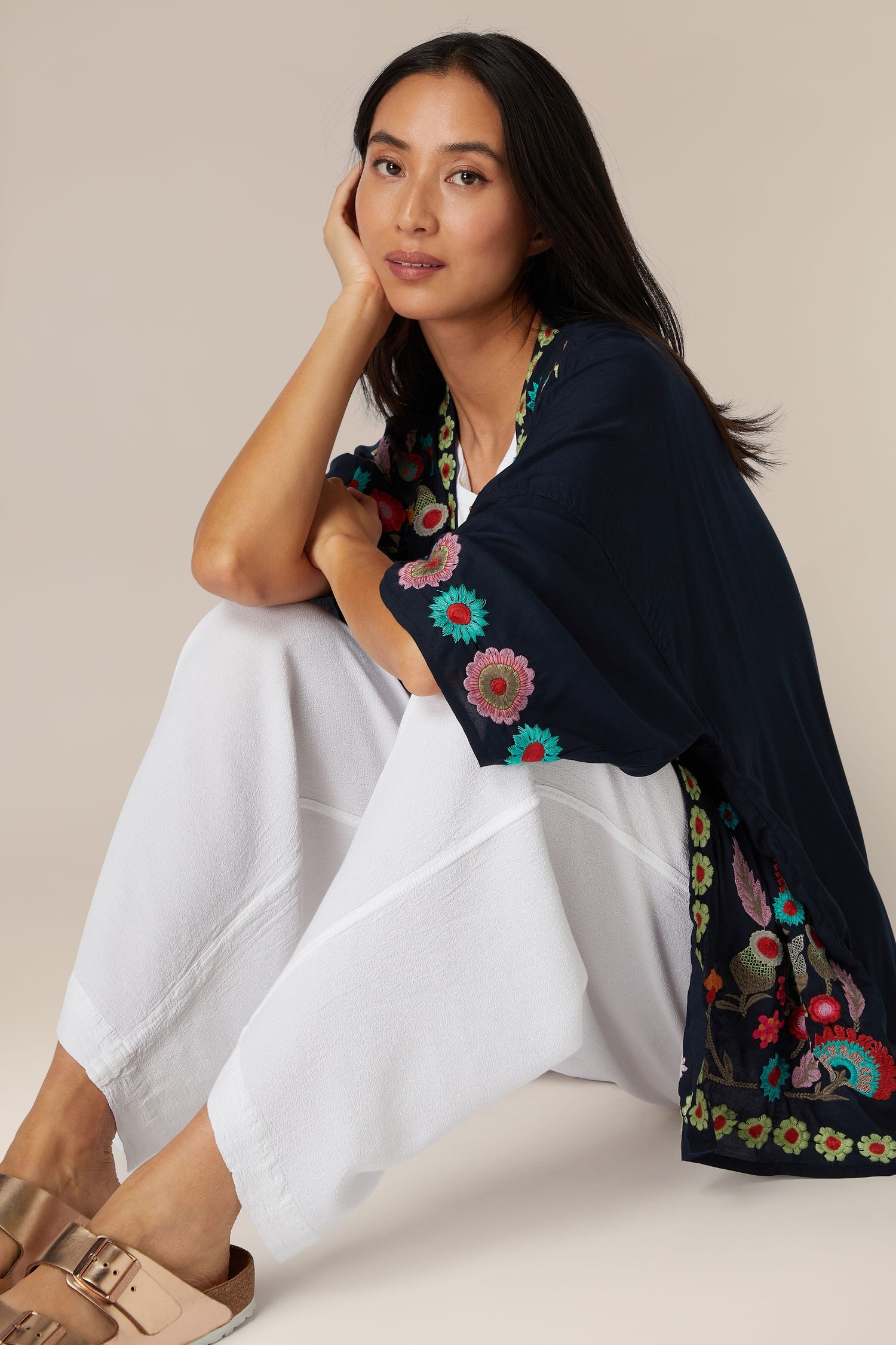 Woman sitting casually in white pants and a navy top adorned with an Embroidered Flower Garden Kimono.