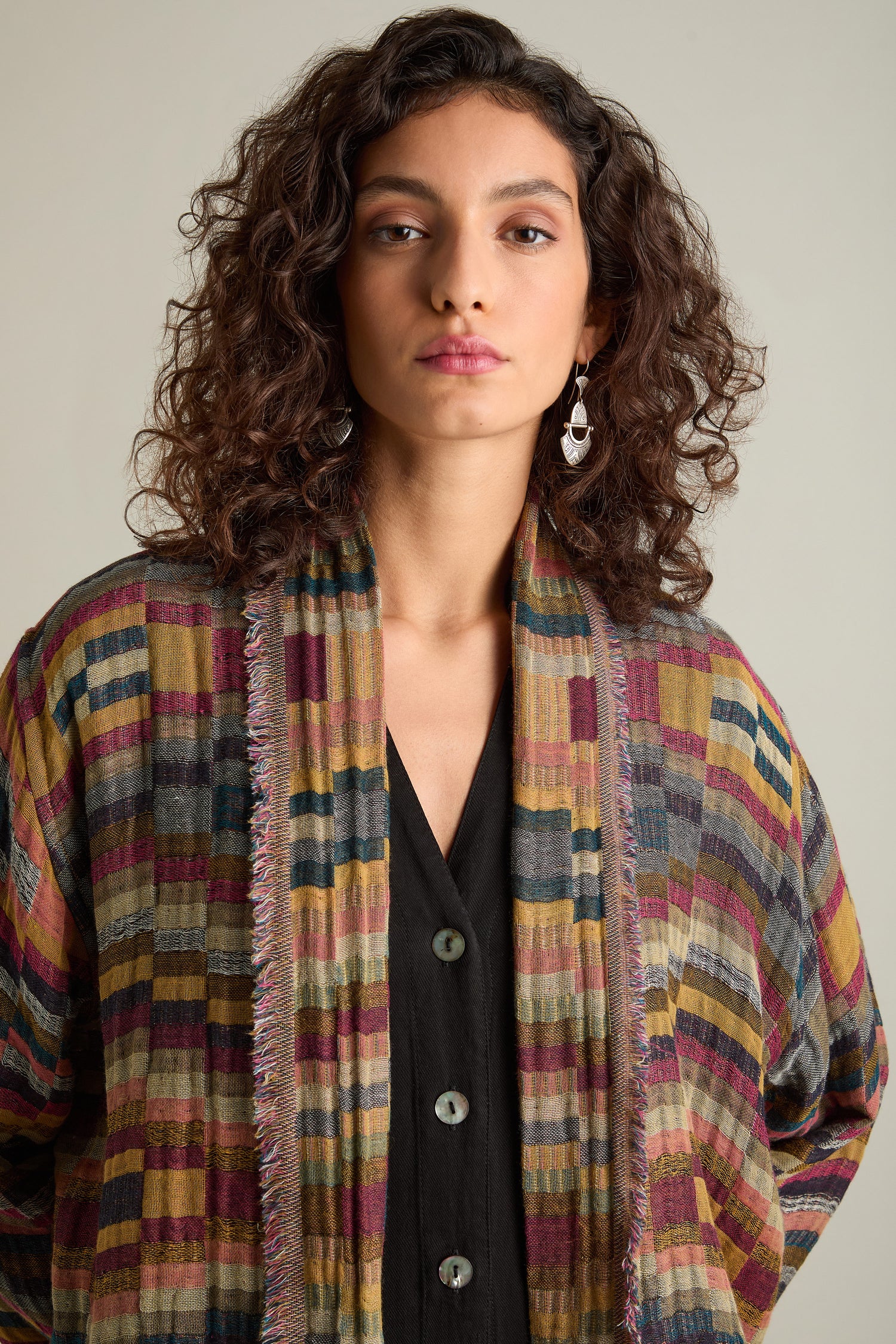 A woman stands facing the camera, wearing a vibrant Hand Stitched Check Jacket over a black top with buttons. She has curly hair and is adorned with earrings that emphasize her timeless style and artisanal craftsmanship.