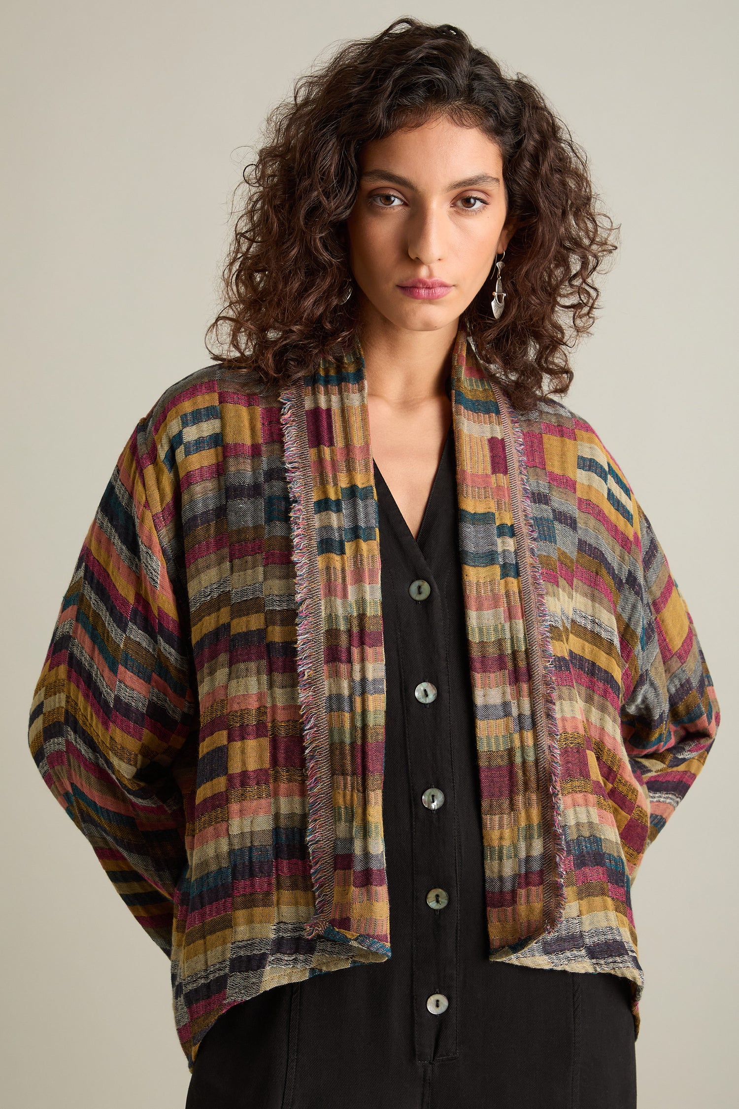 A woman with curly hair wears a vibrant, hand-stitched check jacket over a black button-up shirt, standing against a plain background and looking at the camera. Her timeless style is accentuated by the artisanal craftsmanship of her attire.