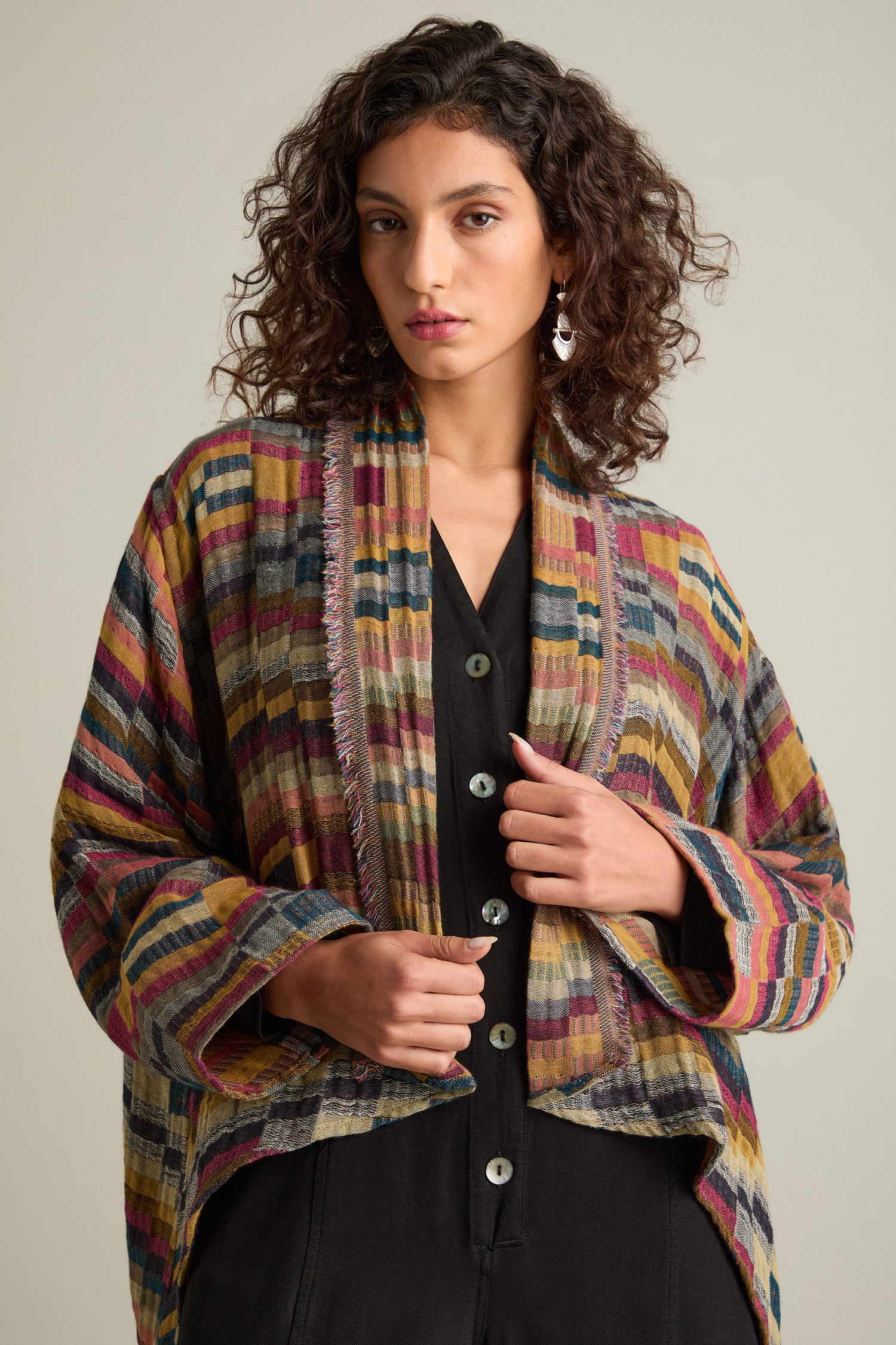 A person with curly hair wears a Hand Stitched Check Jacket over a black button-up top and looks directly at the camera, showcasing timeless style and artisanal craftsmanship.