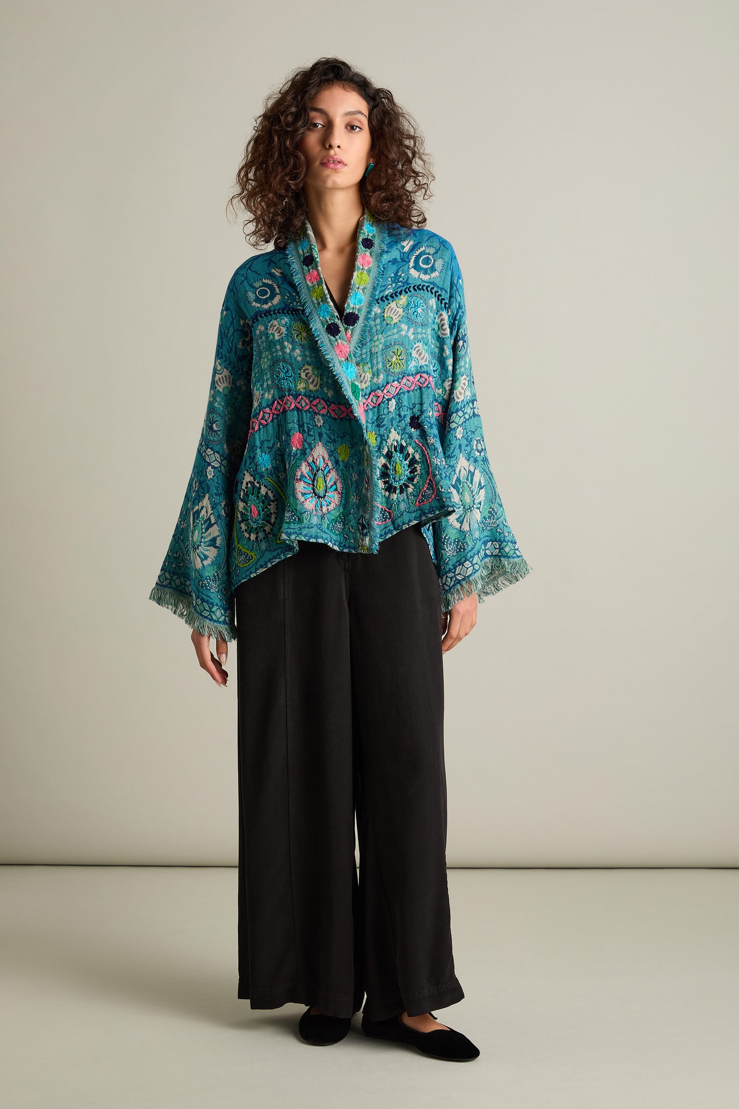 A person with curly hair stands against a plain background, wearing a Cerulean Hand Embroidered Jacket with wide sleeves over a black top and black wide-legged pants.