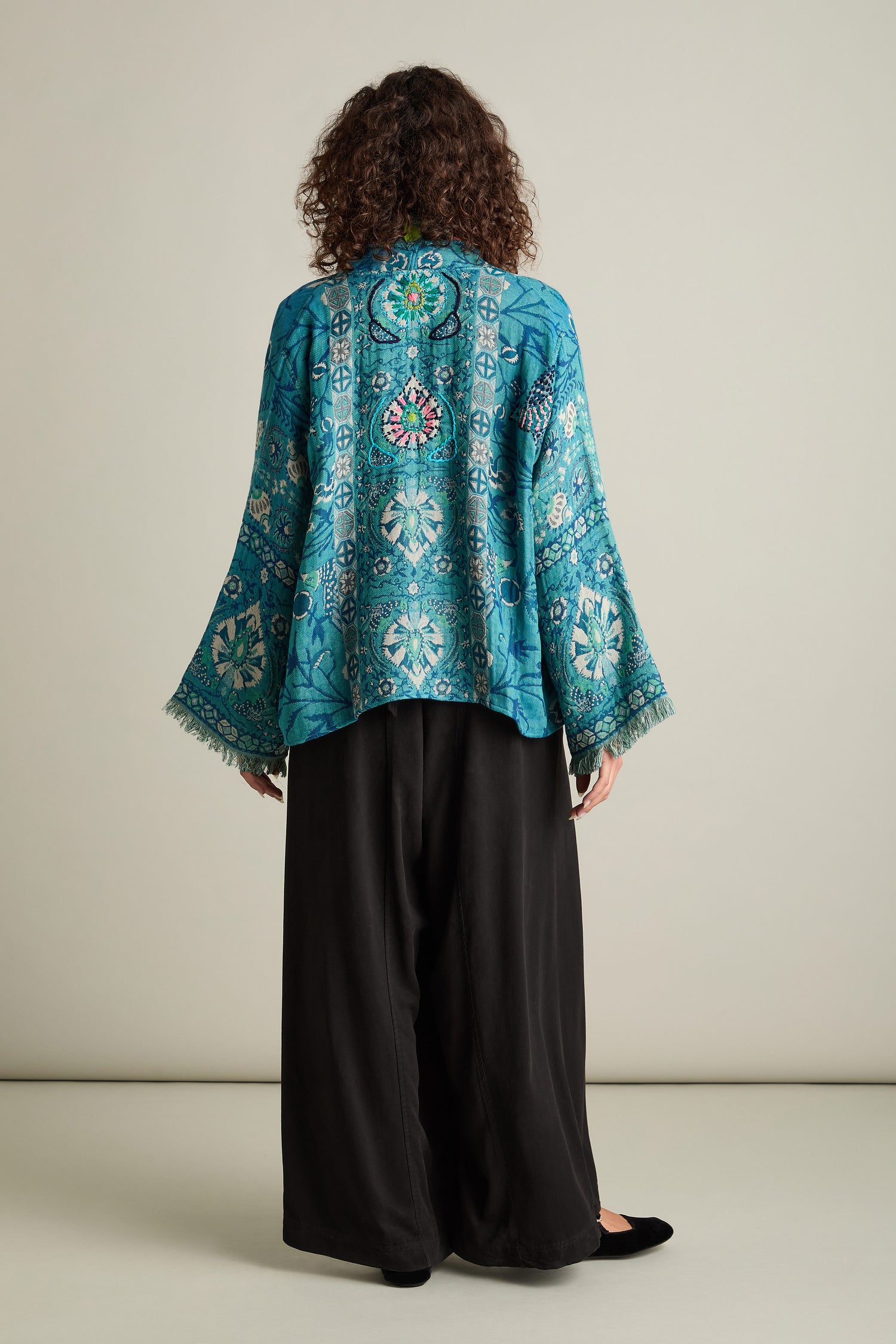 A person with curly hair stands facing away, wearing the Cerulean Hand Embroidered Jacket, which features a kimono-style design with wide sleeves, paired with black loose-fitting pants.