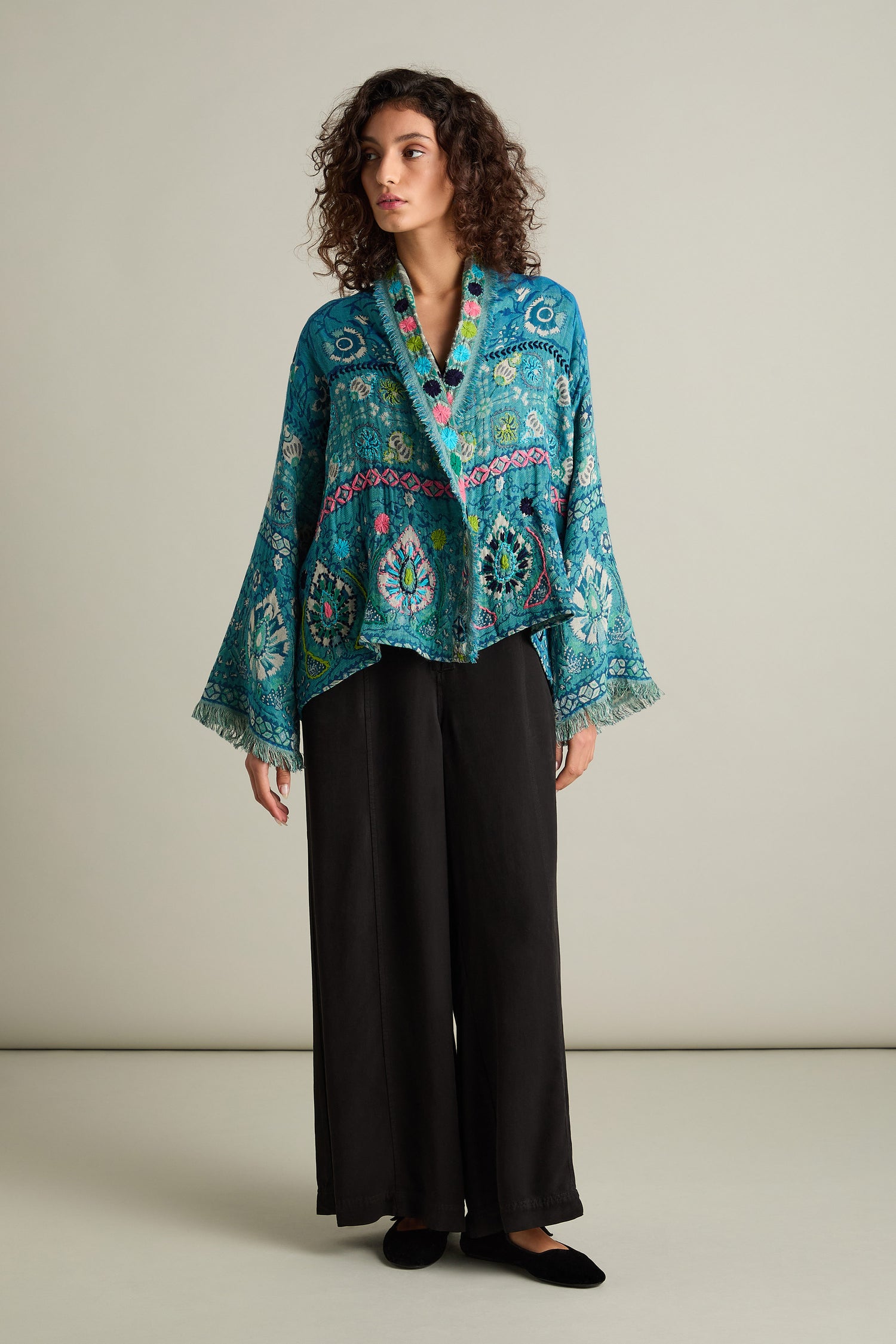 A woman with curly hair is wearing a Cerulean Hand Embroidered Jacket with wide sleeves, paired with black, loose-fitting pants. She stands in front of a plain, light-colored background.