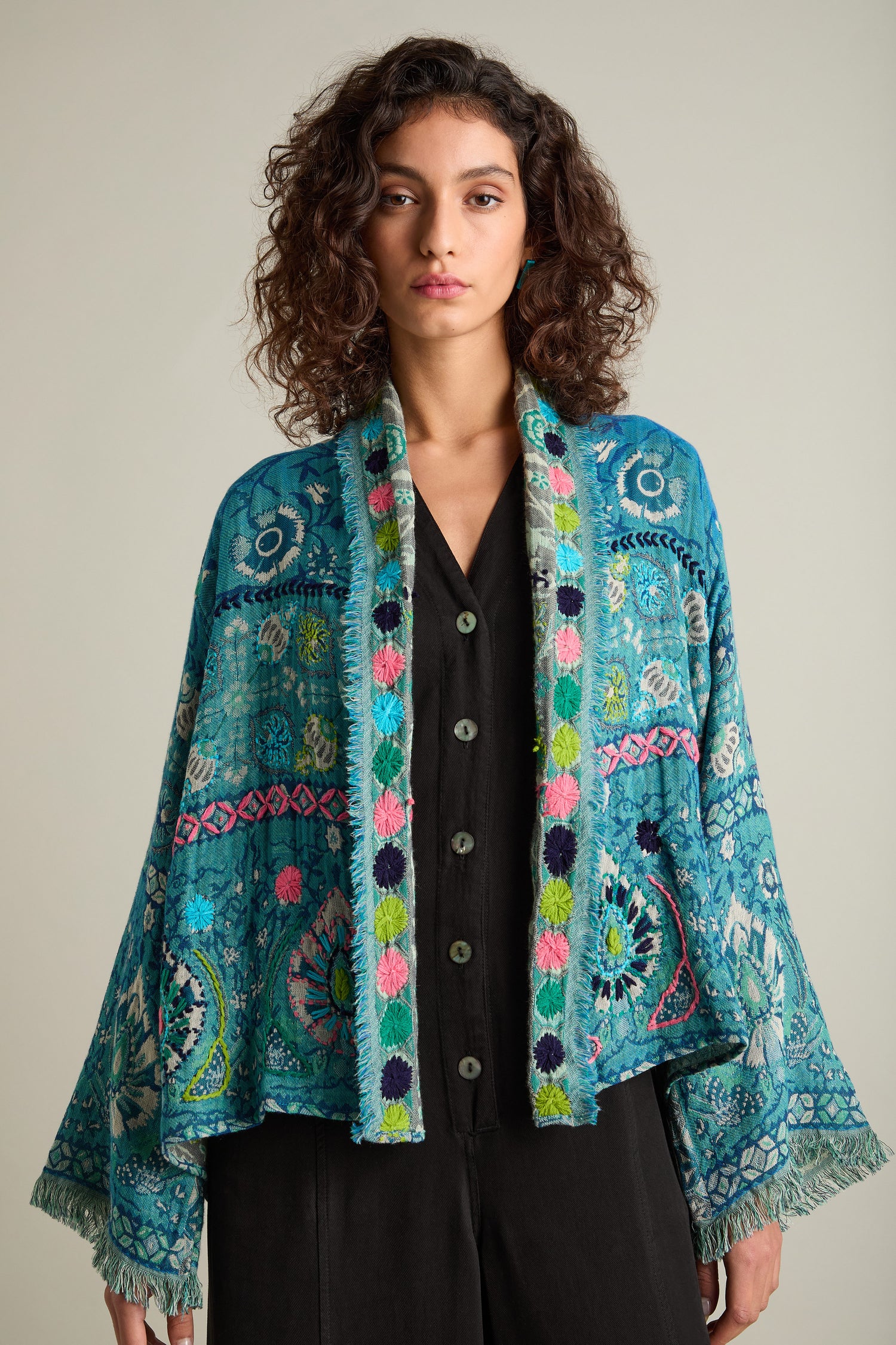 A woman with curly hair is wearing a colorful, patterned Cerulean Hand Embroidered Jacket over a black top. She has a neutral expression and is facing the camera.