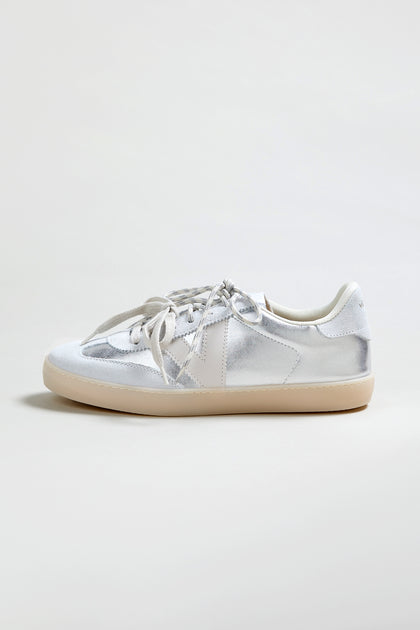 The Berlin Metallic Trainers are silver low-top sneakers with white laces, a white heel tab, and a light beige rubber sole, displayed against a plain light background.