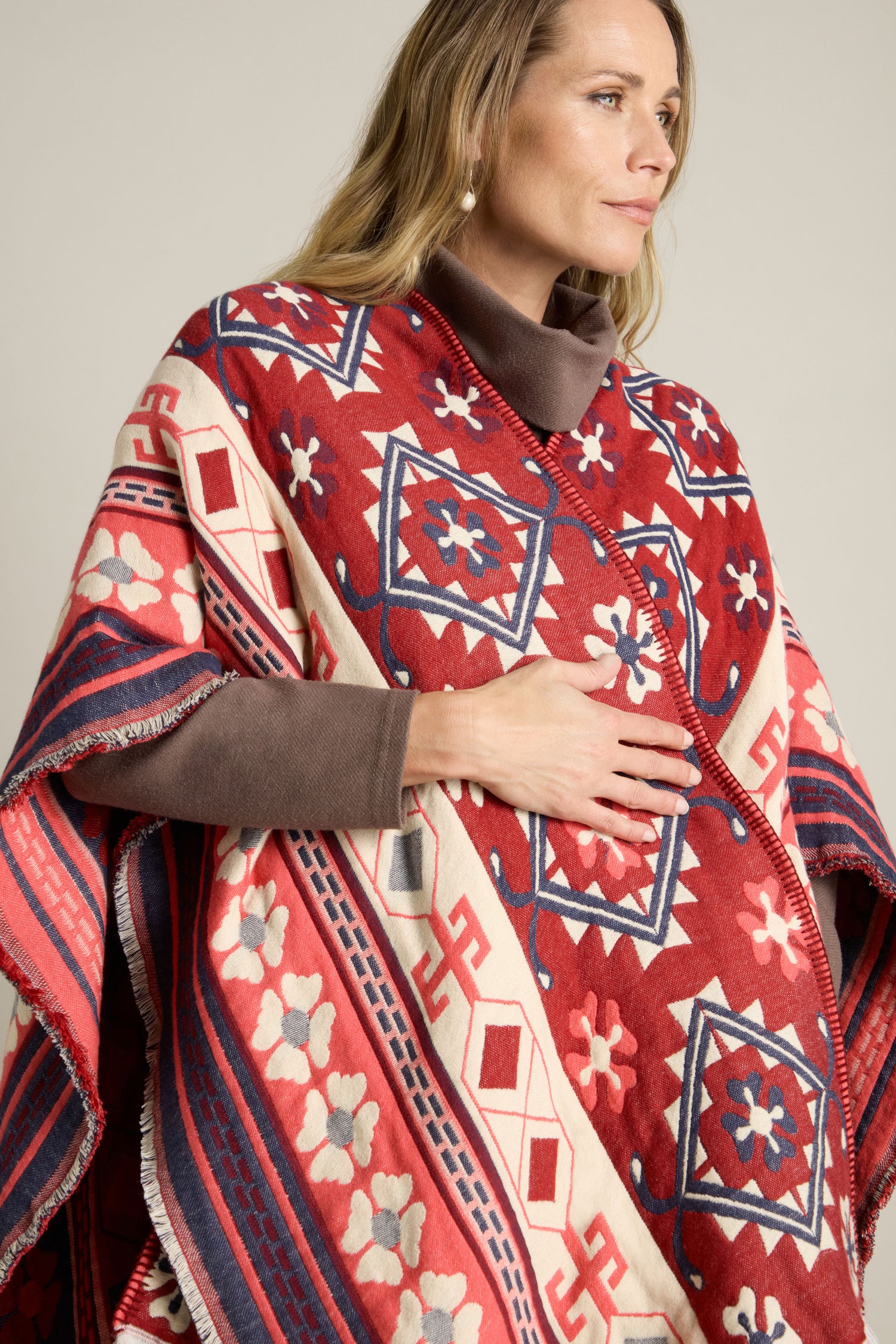 A person standing and looking to the side, wearing an Aztec Woven Shawl in red, blue, and white over a brown long-sleeve top—perfect for an autumn wardrobe.