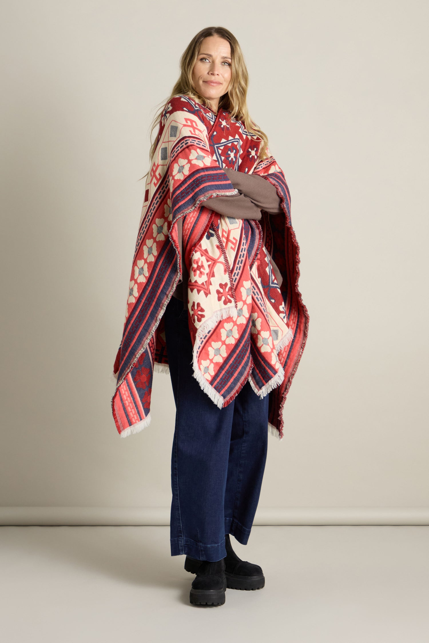 A person stands with arms crossed, showcasing the Aztec Woven Shawl adorned with intricate floral and geometric patterns over a brown top and jeans, making it an ideal addition to an autumn wardrobe.