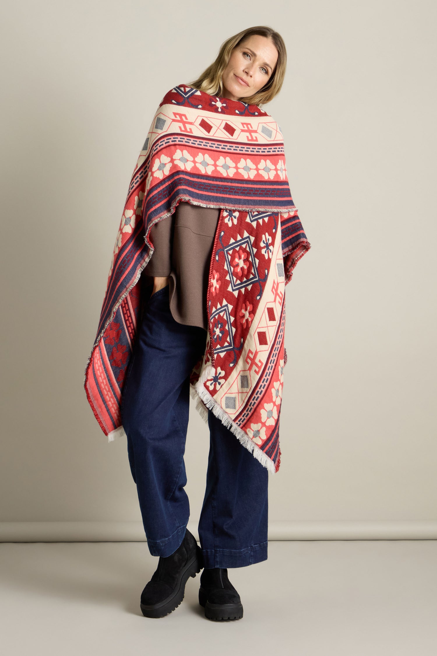 A person stands against a plain background, wrapped in the Aztec Woven Shawl and wearing blue pants and black boots.