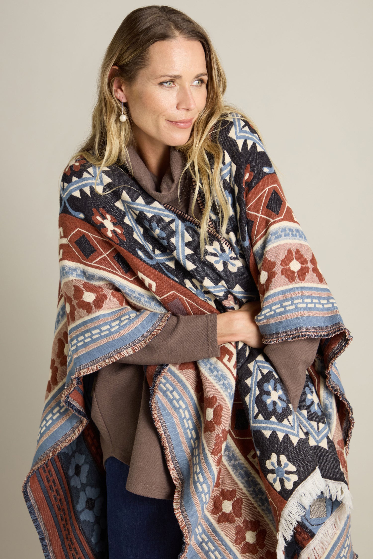 A person with long hair stands against a plain background, adorned in an Aztec Woven Shawl. The shawl showcases intricate geometric designs in shades of blue, brown, and white, making it the perfect addition to any autumn wardrobe.