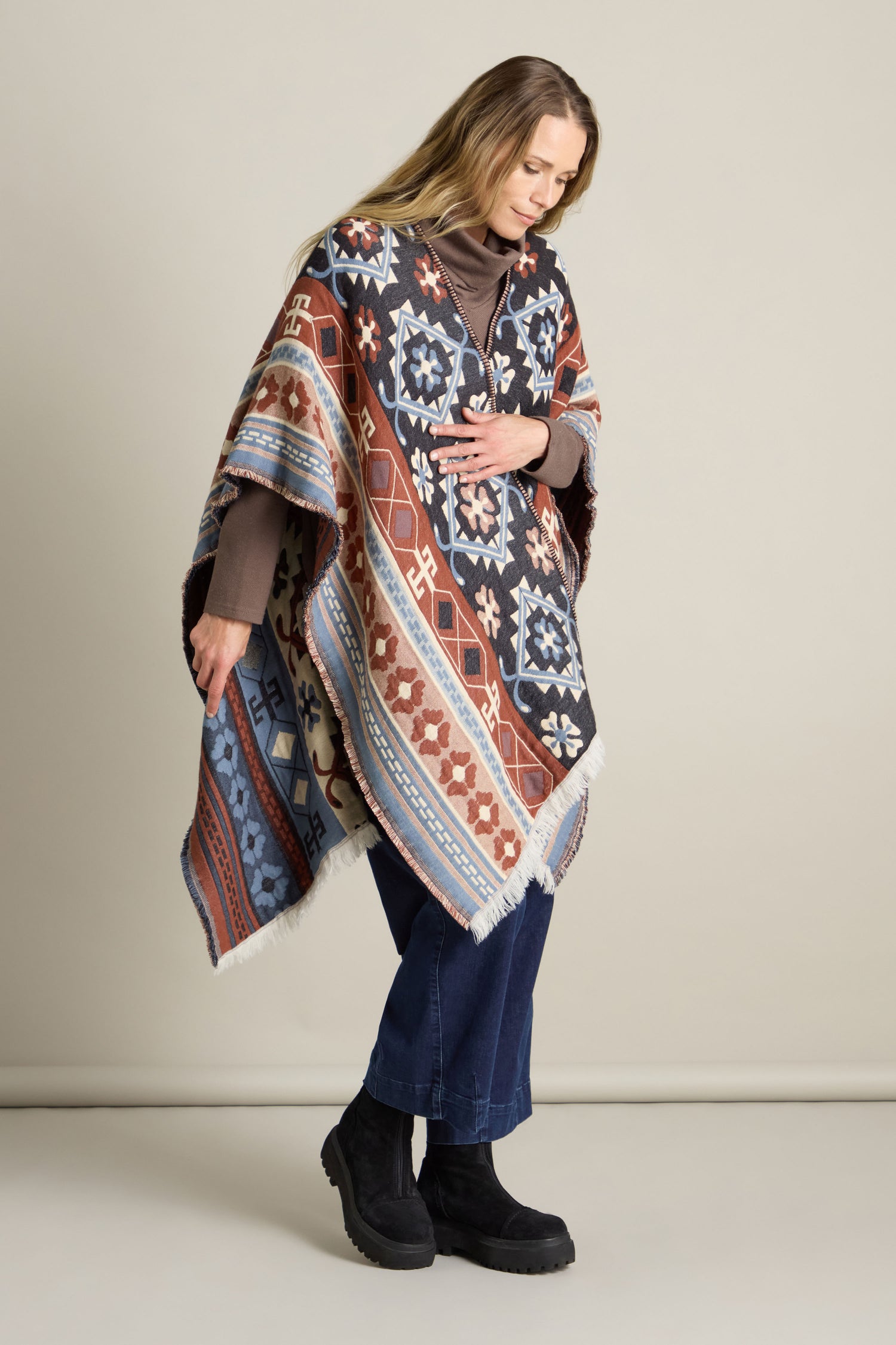 A person stands wearing an Aztec Woven Shawl with a multicolored pattern, a brown top, blue wide-leg pants, and black boots against a plain background.