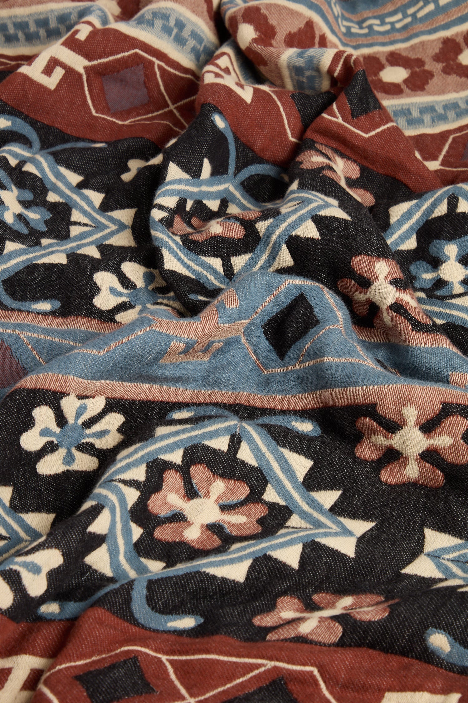 The Aztec Woven Shawl showcases an intricate floral and geometric pattern in black, blue, red, and beige colors, beautifully draped with folds and shadows, making it perfect for your autumn wardrobe.