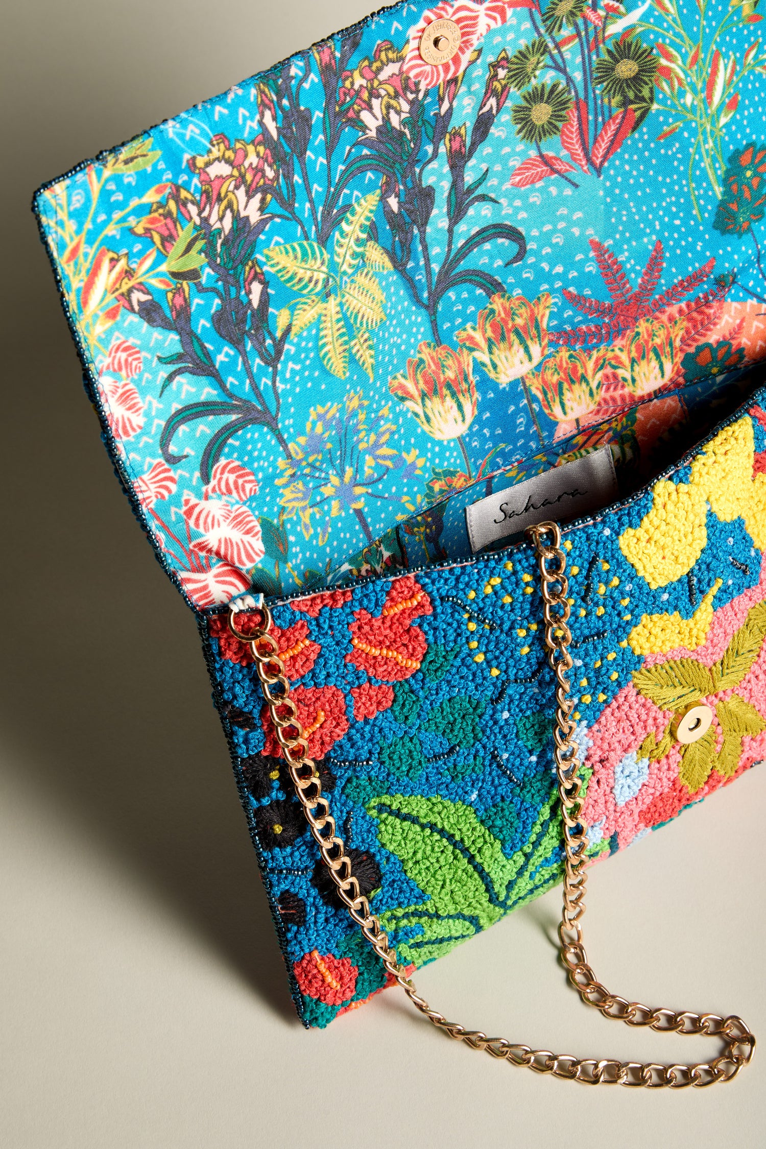 The Full Bloom Hand Beaded Clutch Bag exudes a vibrant floral beauty with its colorful embroidery and gold chain strap, opening to reveal a lively floral-patterned interior.