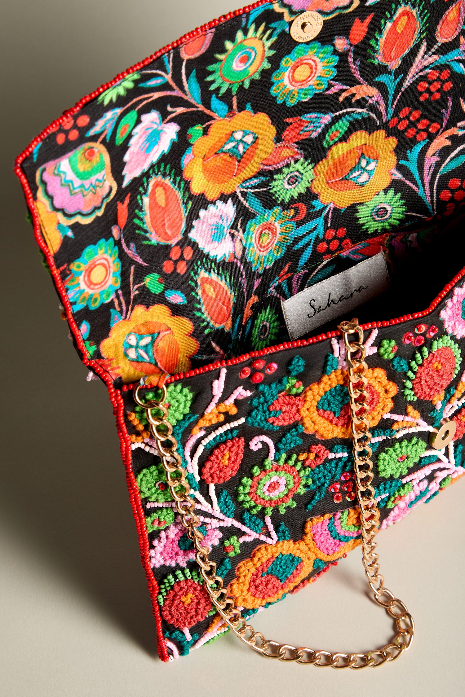 The Wild Garden Hand Beaded Clutch Bag showcases a vibrant floral beaded design in its colorful embroidery. Complete with a gold chain strap and red beaded trim, the interior fabric harmonizes with the vivid floral pattern. This bold accessory is ideal for enhancing any outfit with flair.