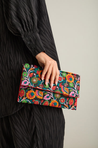 A person holding a Wild Garden Hand Beaded Clutch Bag, its floral design contrasting beautifully with their long-sleeved black garment, creating a striking statement accessory.