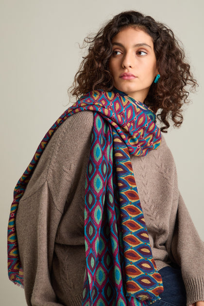 A person with curly hair, dressed in a brown sweater and adorned with a Retro Wool Twill Scarf that boasts a colorful patterned design, gazes off to the side against an unadorned background.